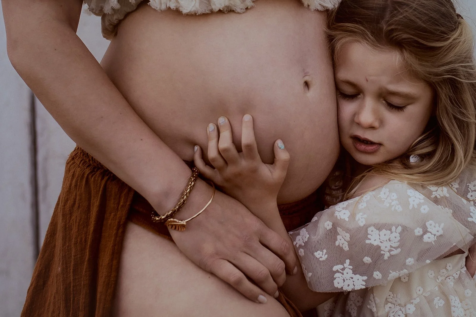 A pregnant woman with her hand on her stomach and a young girl hugging her belly tightly, with the girl resting her head on the woman's stomach, in a tender moment.