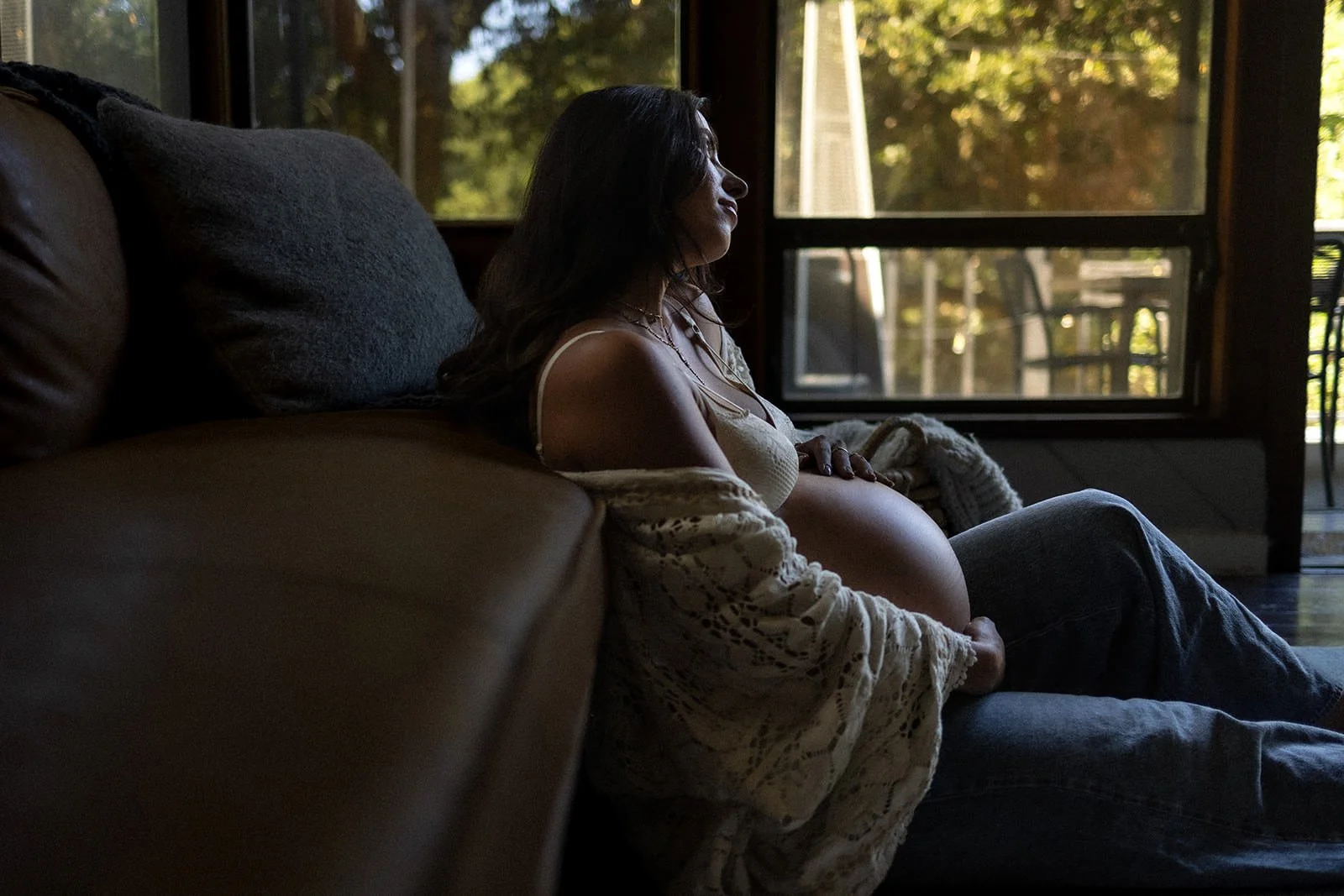 A pregnant woman sitting on a couch near a large window, with her eyes closed, in a cozy, warmly lit room with trees visible outside.