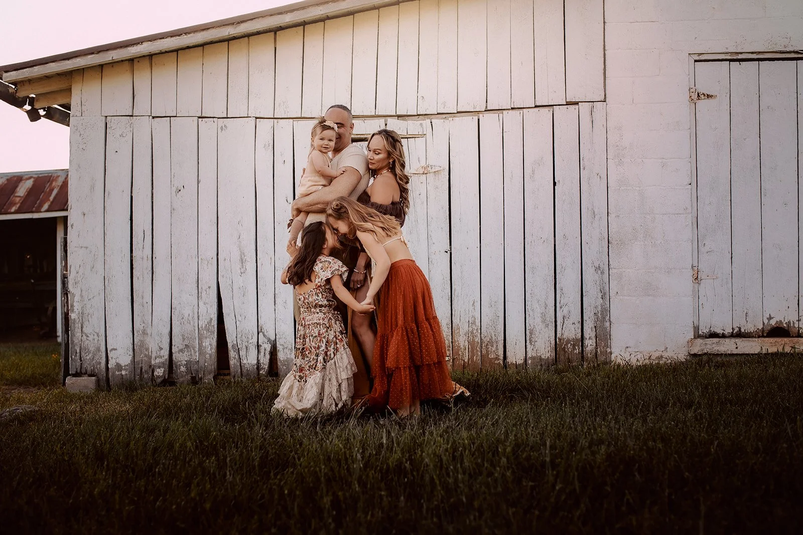A group of five women, including children, are standing and hugging outside in front of a weathered white barn at sunset.
