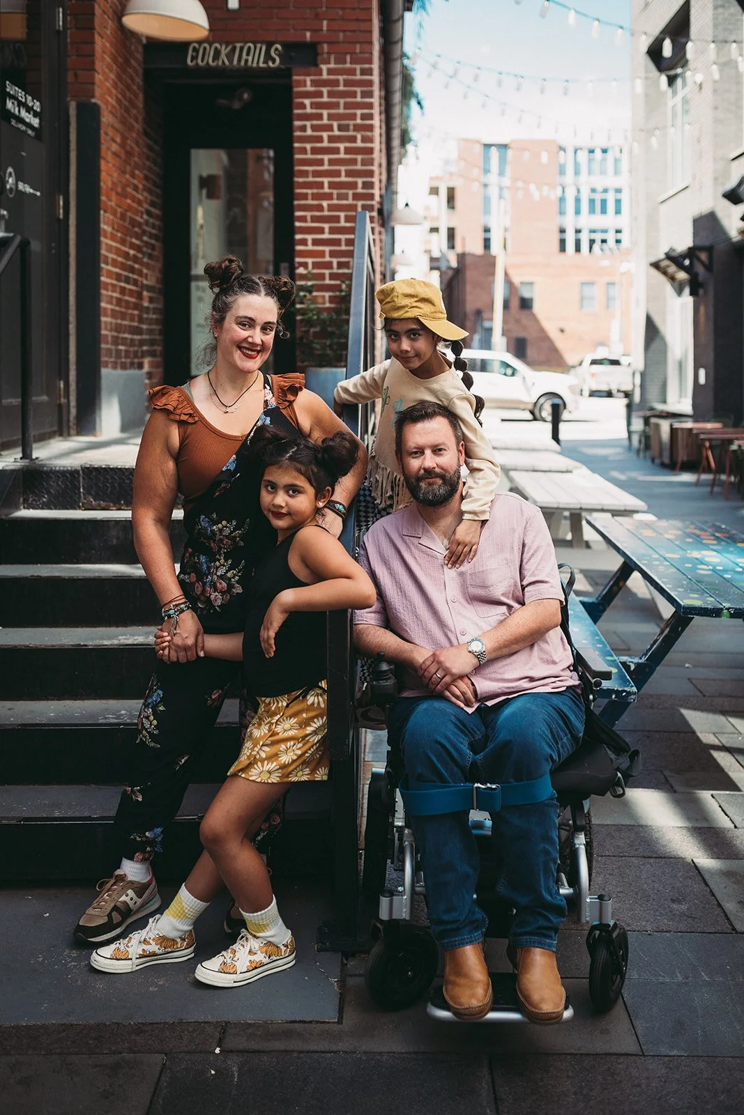 A diverse family of four, including a man in a wheelchair, two young girls, and a woman, posing on an outdoor urban patio with brick walls, string lights, and tables.