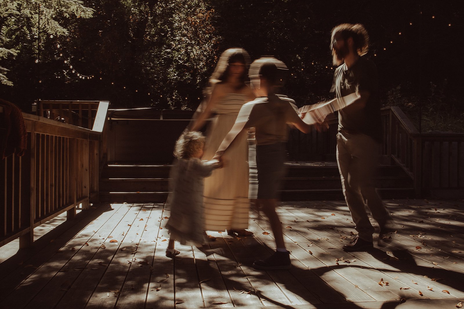 Family dancing on a wooden deck at night, with string lights and trees in the background, some motion blur in the photo.