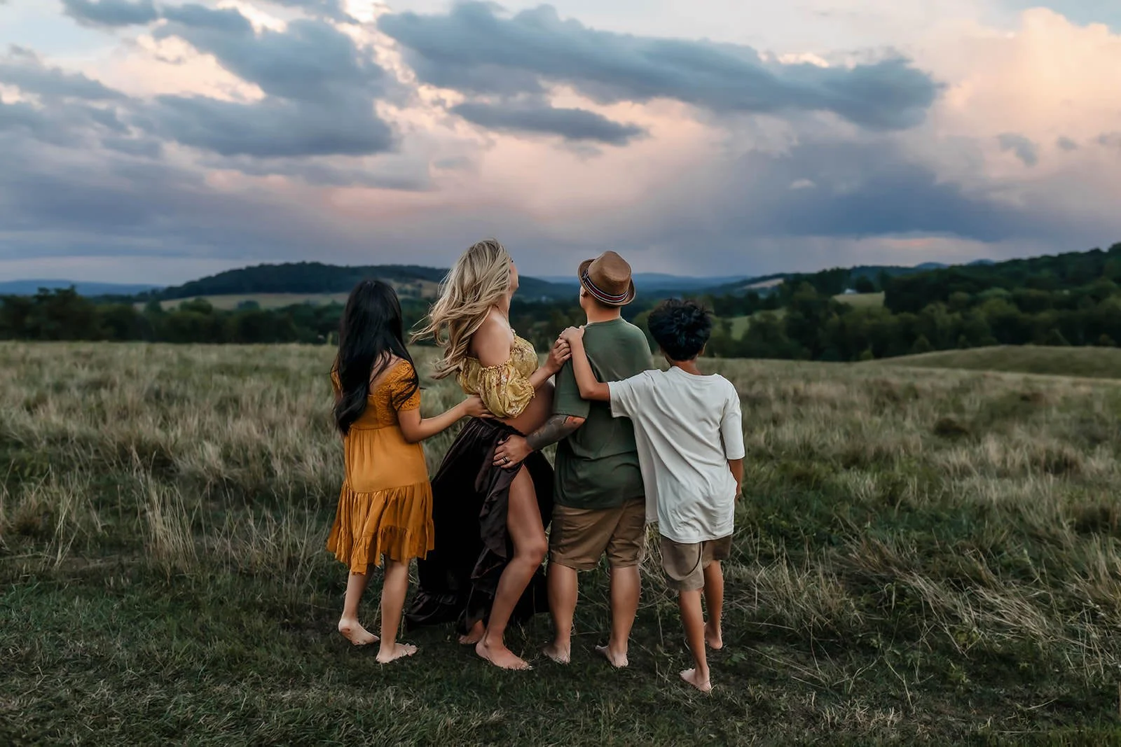A group of five diverse people standing in a grassy field with rolling hills and cloudy sky in the background; two adults, three children, everyone embracing or holding hands, looking into the distance.