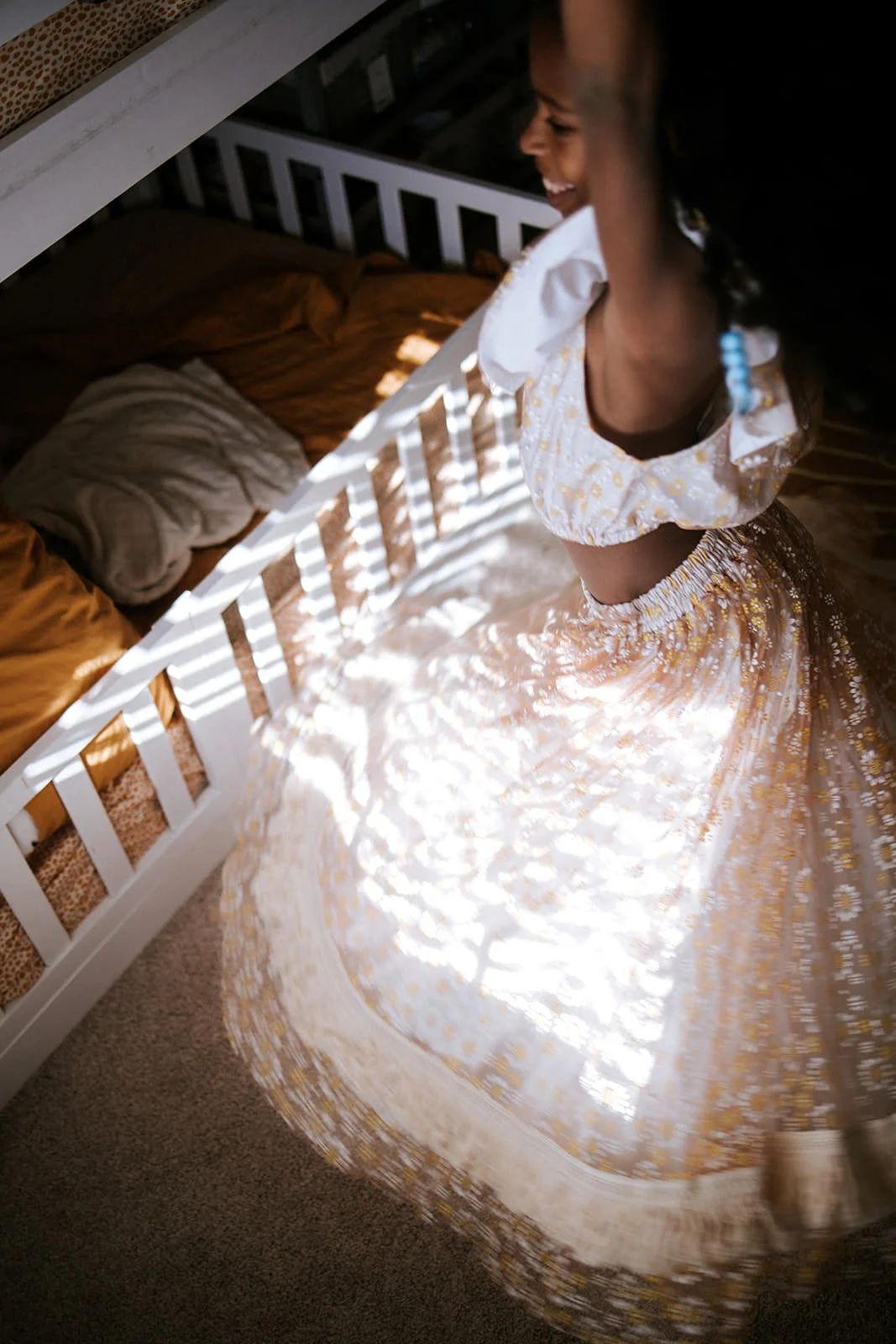 A young girl in a yellow and white floral dress spinning happily in a bedroom with sunlight casting shadows through window blinds.
