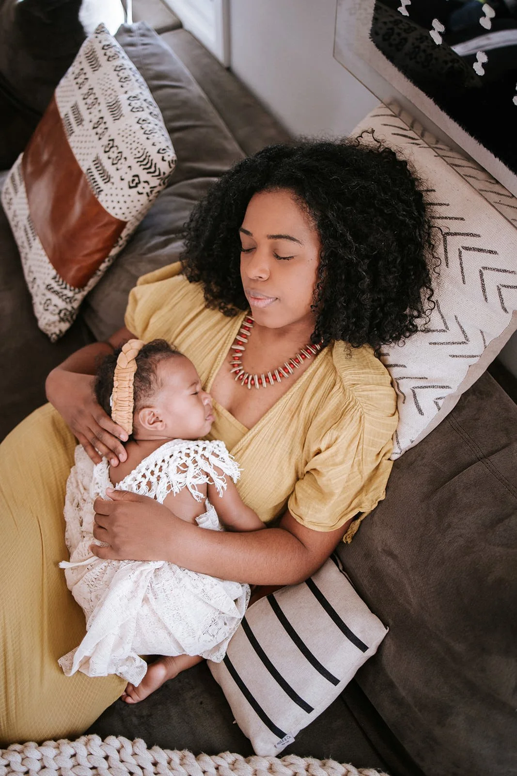 A woman with black curly hair, wearing a yellow dress and a red necklace, lies on a couch with her eyes closed, holding a sleeping baby dressed in white with a lace dress and a beige headband.