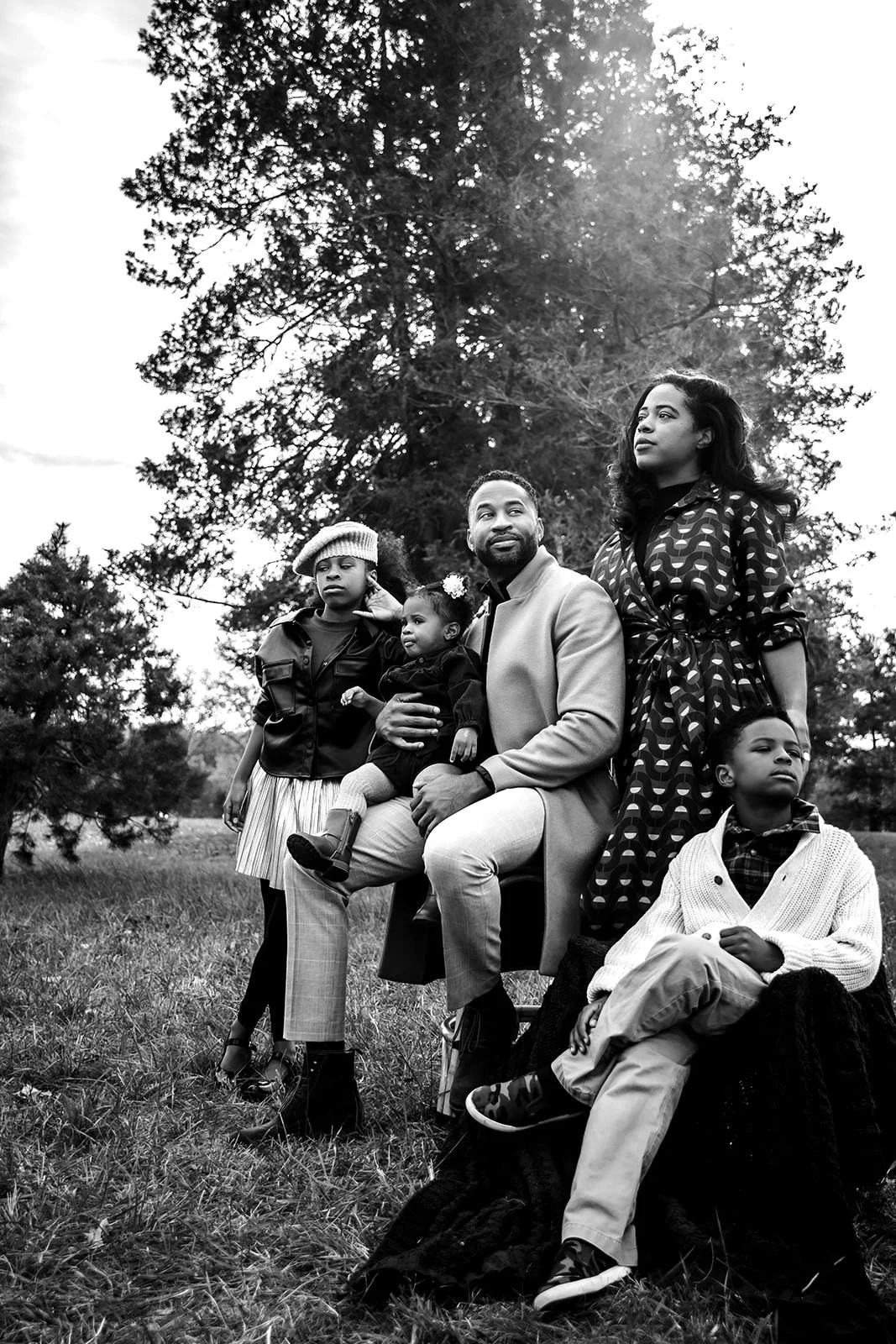Black and white photo of a family of five outdoors near trees, with a man sitting and holding a young girl, a woman standing nearby, and a boy and girl sitting on the grass.