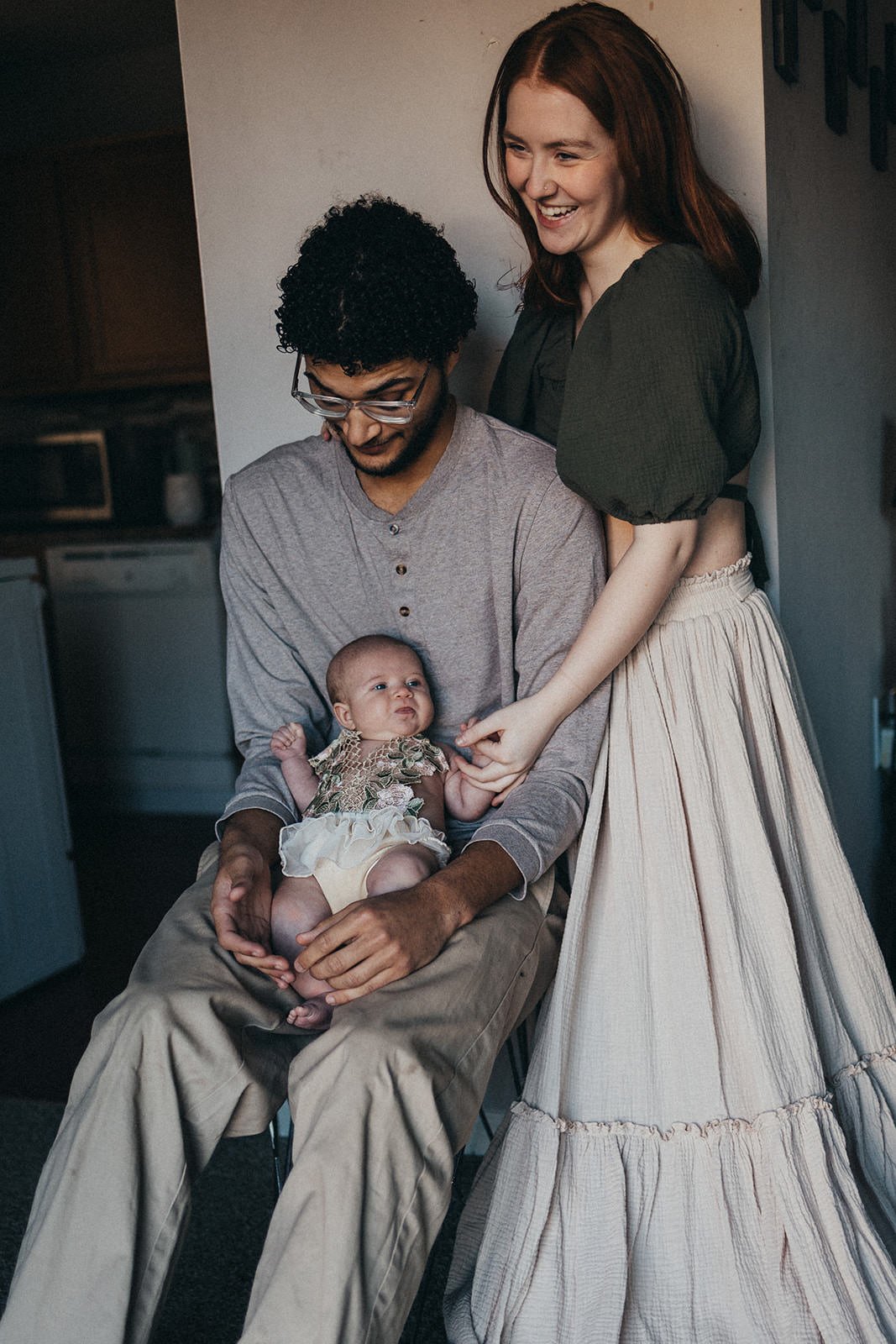 A woman and a man sitting in a room with a baby, while another woman stands beside them holding hands with the baby. The woman is smiling and the man is looking down at the baby, who is sitting on the man's lap.