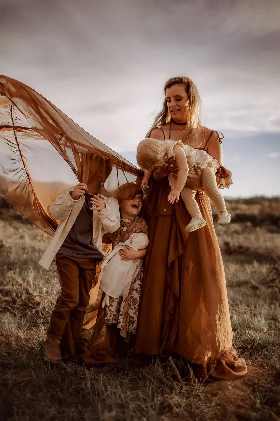 A woman surrounded by three children outdoors on a grassy field, with two children partially hidden under a brown sheer fabric, one girl holding a doll, and a woman holding a small girl in her arms during sunset or sunrise.