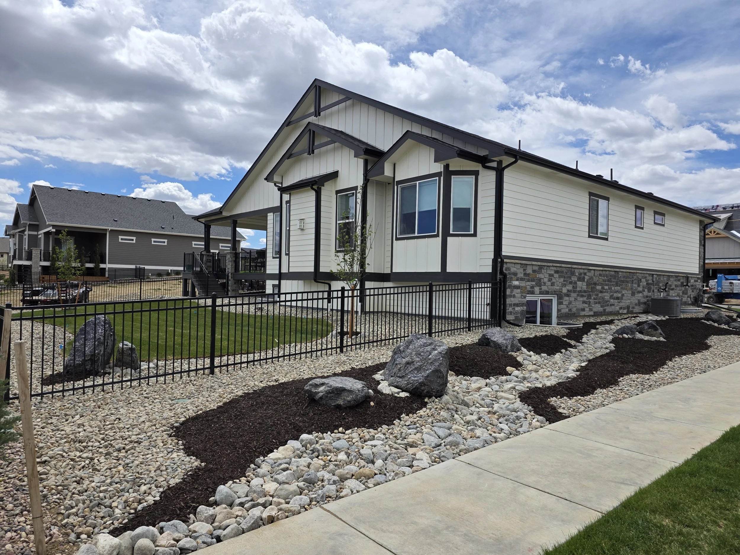 Front yard with rock landscaping, young tree, black metal fence, sidewalk, and white house with black trim under a partly cloudy sky. Big granite boulders, hardscape, dry creek bed