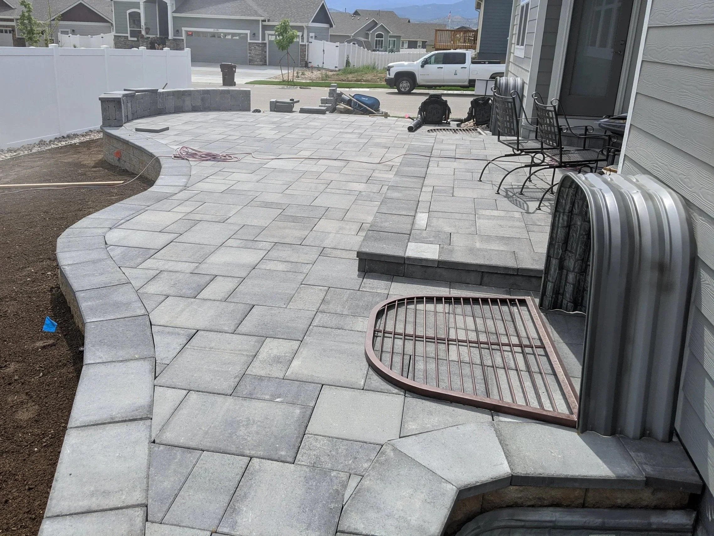 A backyard patio area under construction with pavers, some stacked chairs, and construction tools. A white fence encloses the yard, and neighboring houses are visible in the background. Hardscape, courtyard, paver patio, masonry, Bench
