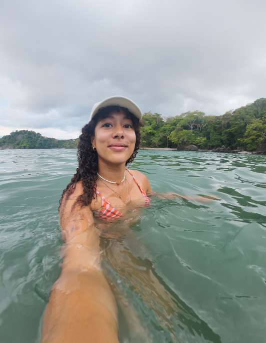 Young woman in a checkered bikini and white cap taking a selfie while swimming in a river with lush greenery and cloudy sky in the background.