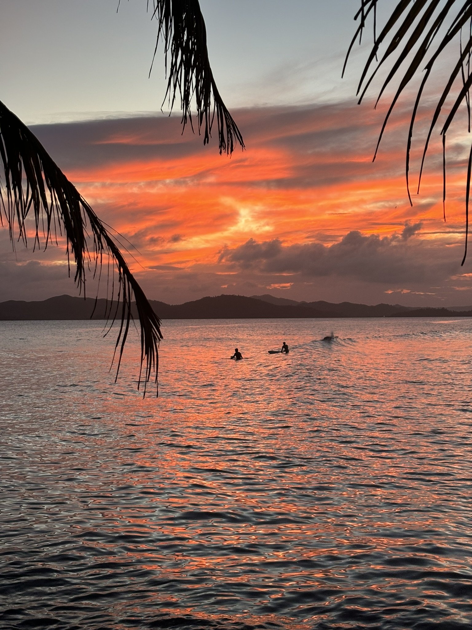 Sunset over a body of water with palm tree leaves in the foreground and mountains in the distance. Two people are visible on the water, possibly surfing or swimming.