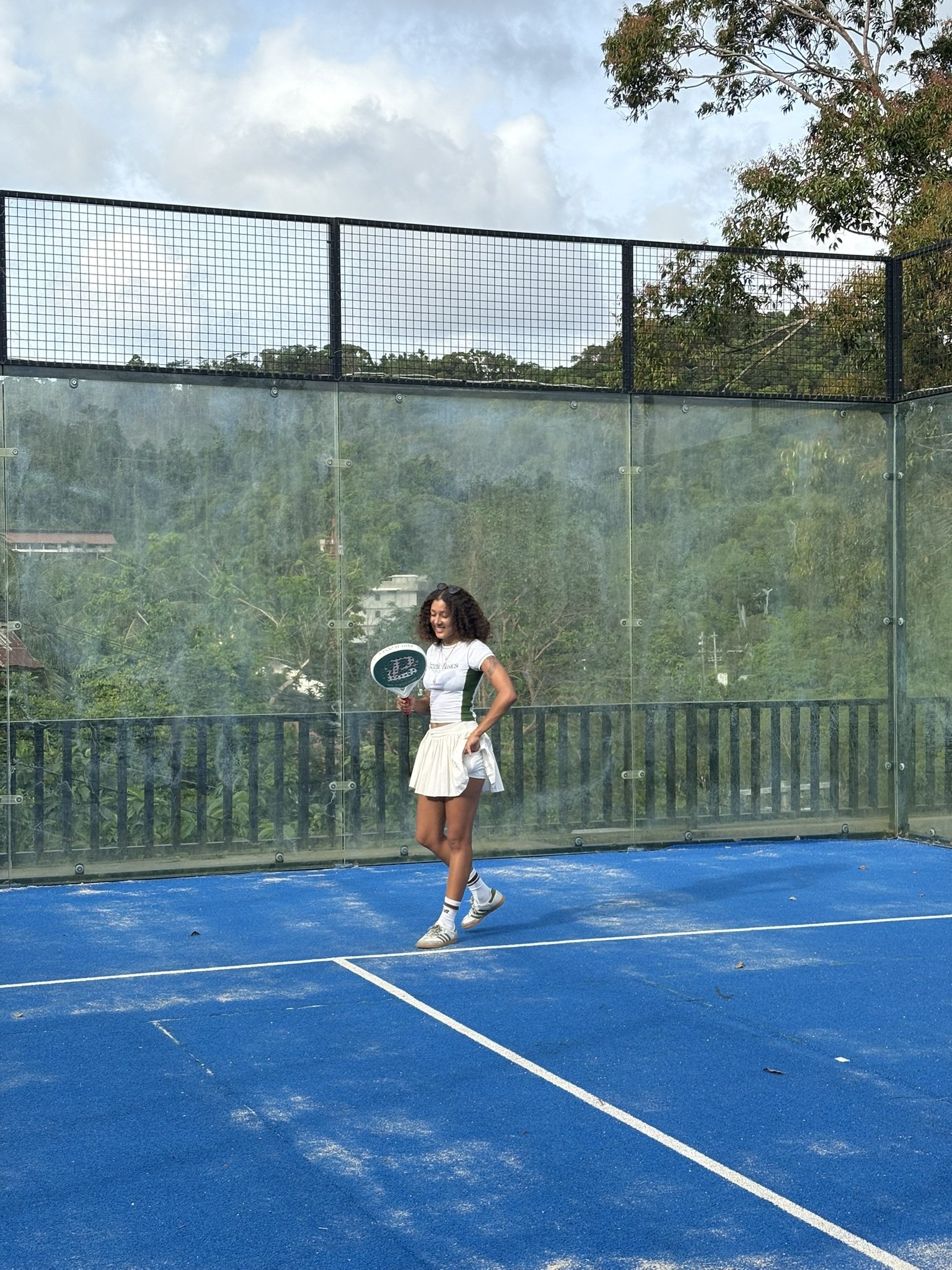A woman with curly hair wearing a tennis outfit and sneakers, holding a badminton racket, standing on a blue tennis court with trees and a partly cloudy sky in the background.