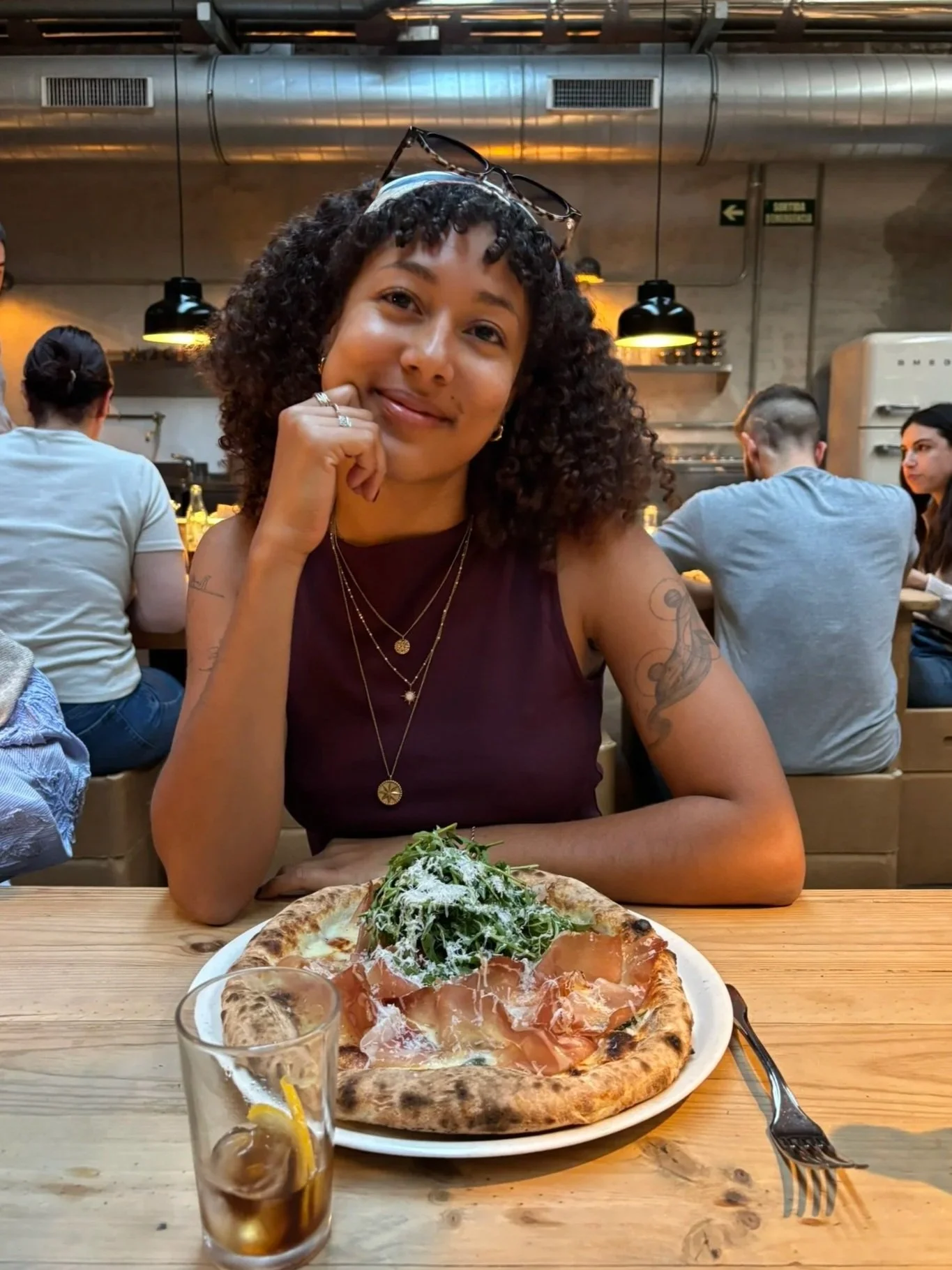 A woman sitting at a wooden table in a restaurant, smiling with her hand resting on her chin. She has curly hair, wears glasses on her head, and layered necklaces. In front of her is a pizza topped with greens, prosciutto, and cheese, along with a glass of iced tea with lemon and a straw. The background reveals other diners and industrial-style lighting.