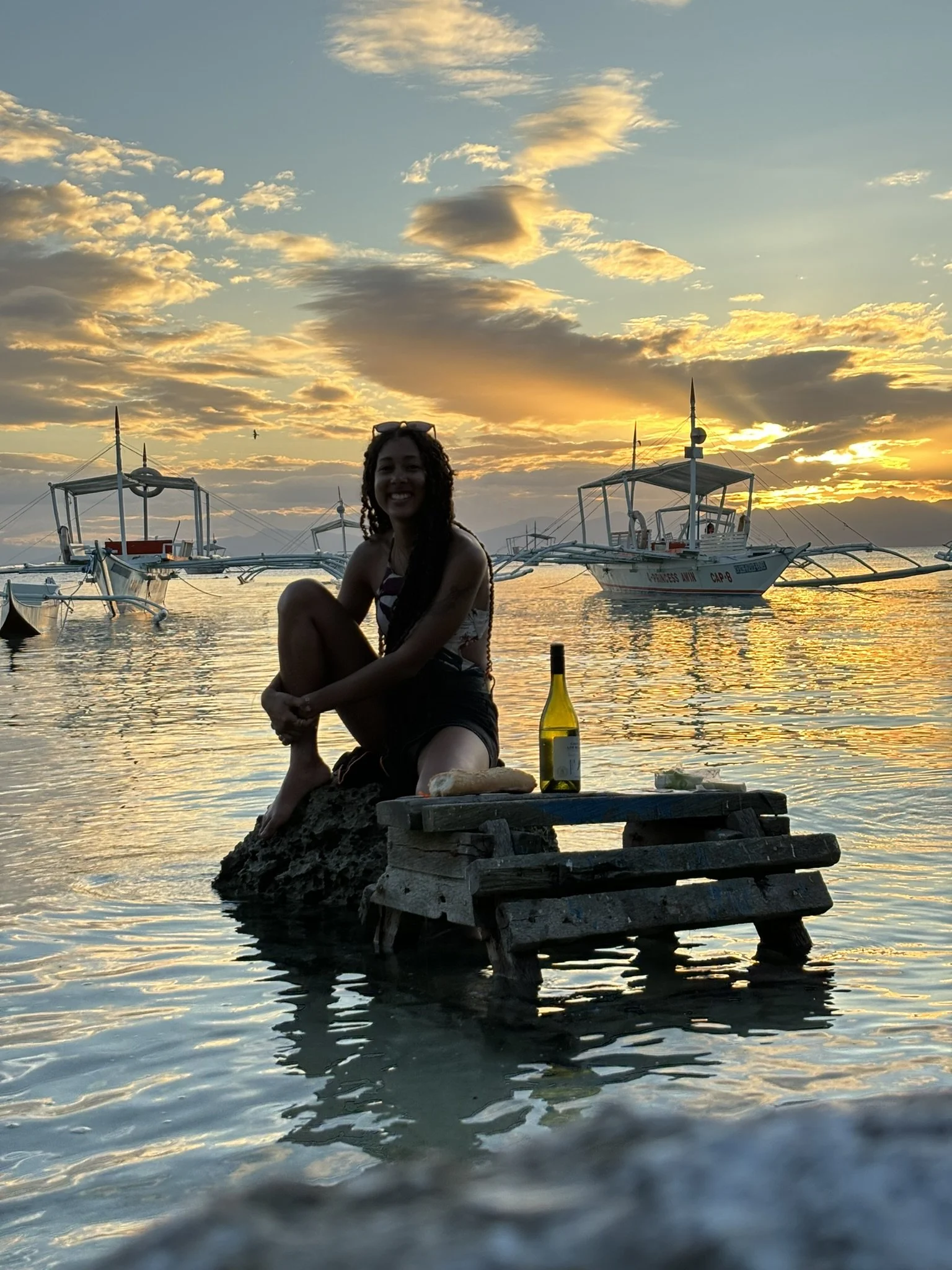 A woman sitting on rocks at sunset near the water, with boats in the background and a bottle of white wine on a small wooden table.