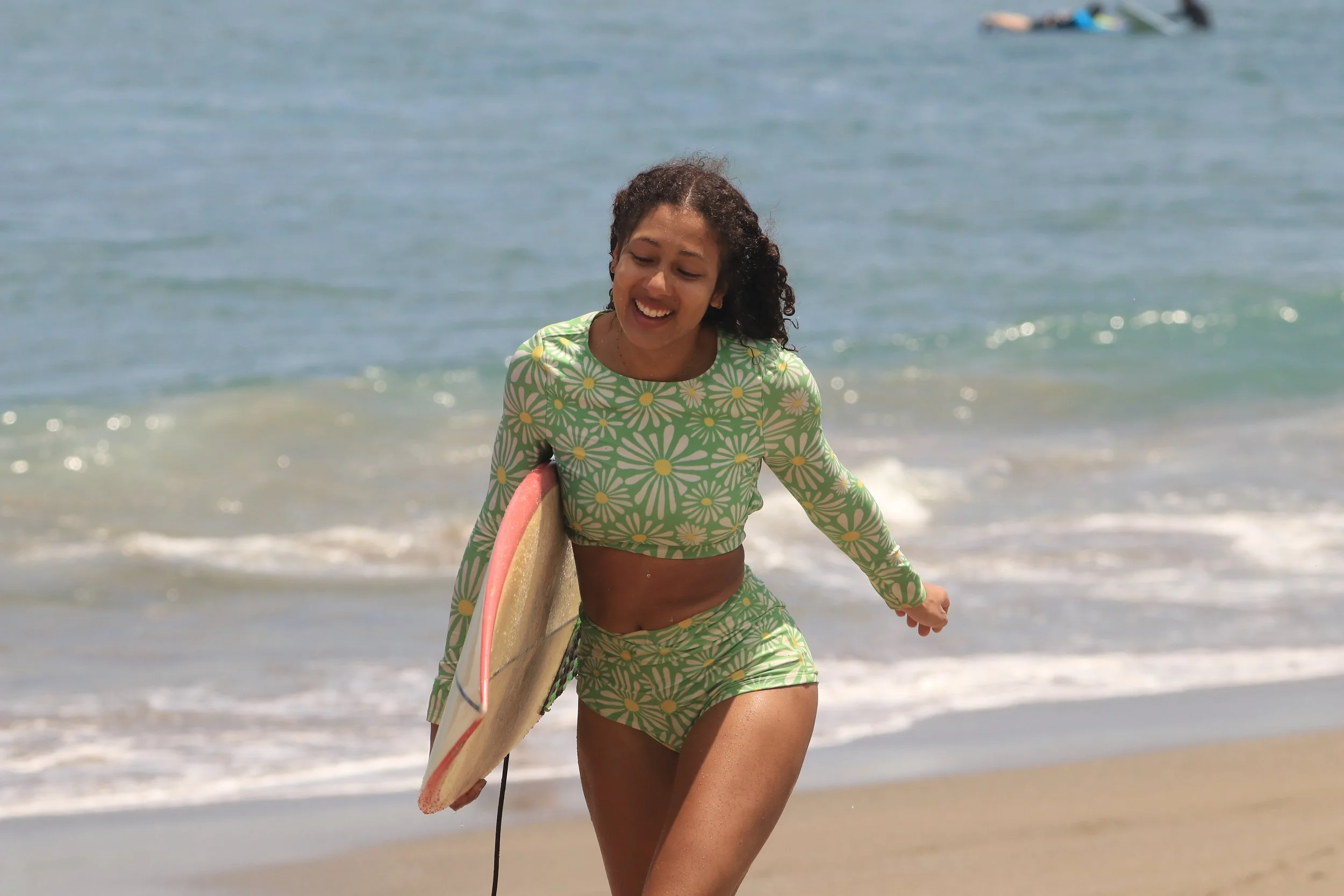 A young woman with curly hair smiling and walking out of the ocean holding a surfboard, wearing a green floral swimsuit.