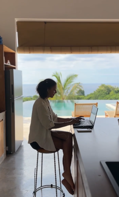 Woman working on laptop at kitchen island with ocean view and palm trees outside.