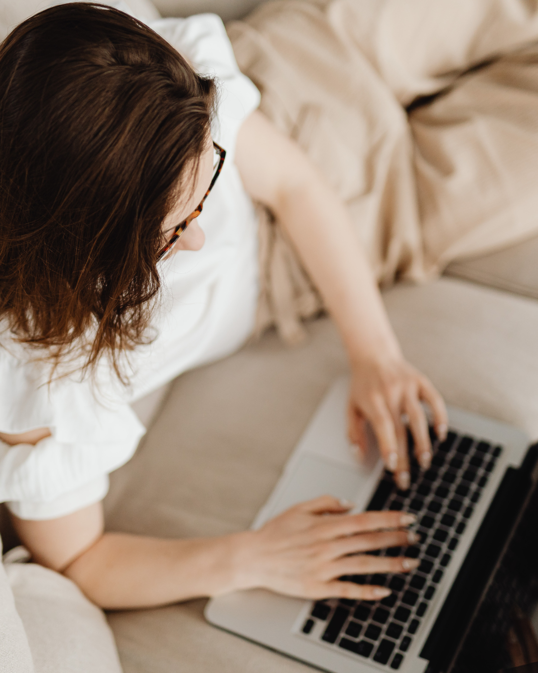 A woman with brown hair and glasses is lying on a beige couch, using a laptop.