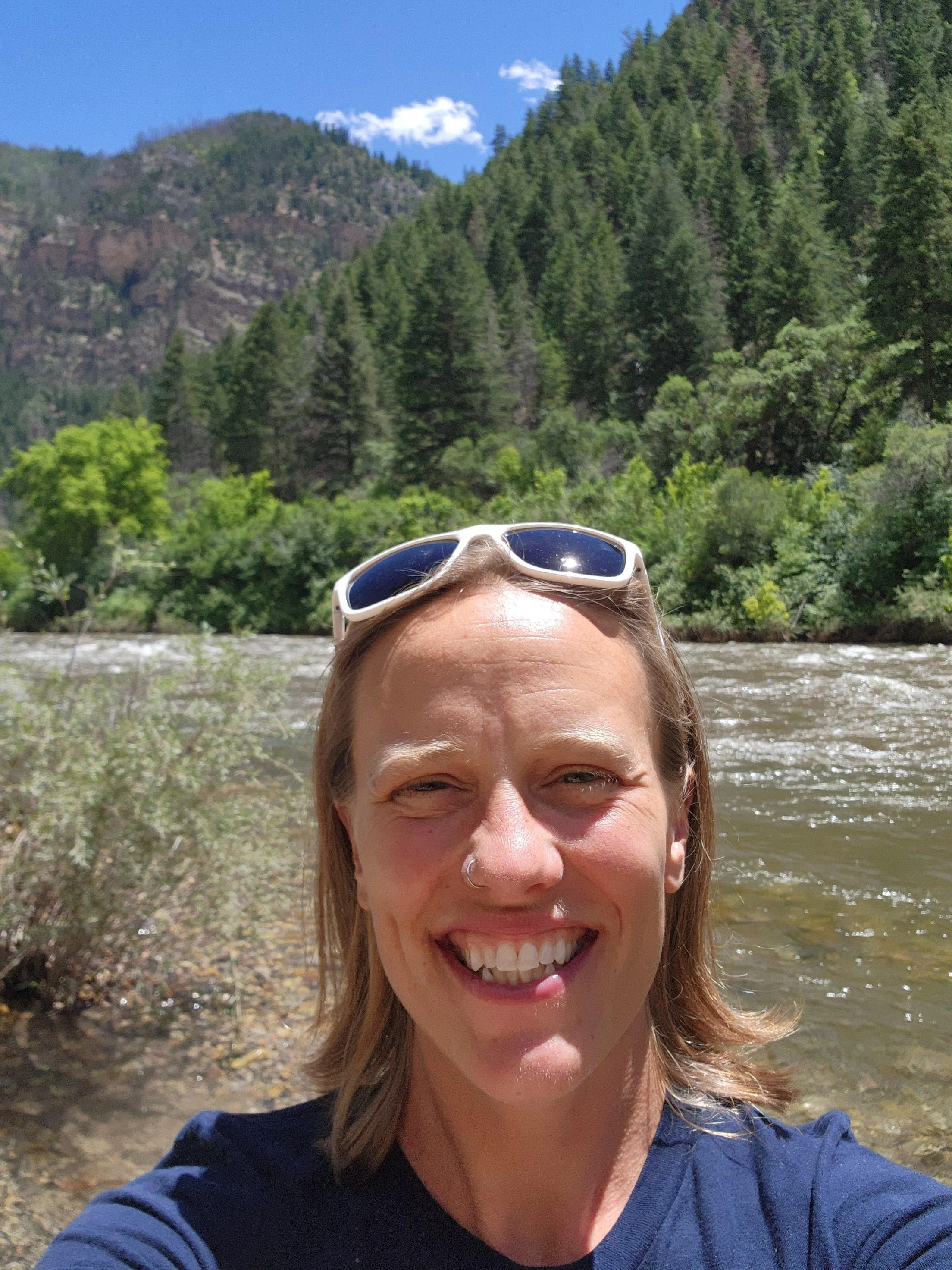 A woman with sunglasses on her head smiling near a river with a forested mountain in the background under a blue sky with some clouds.