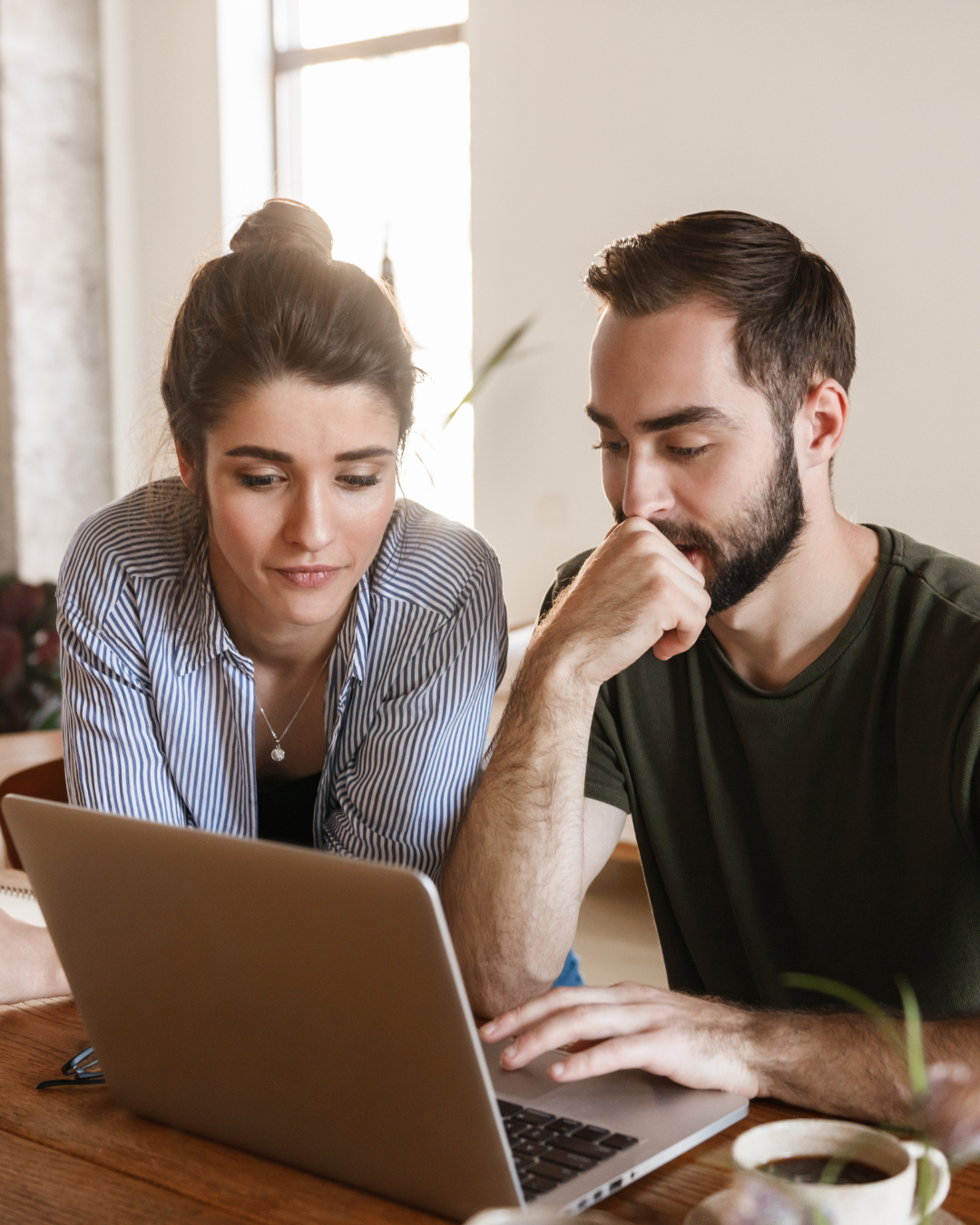 Two people looking at a laptop screen together, sitting at a wooden table, in a bright room.