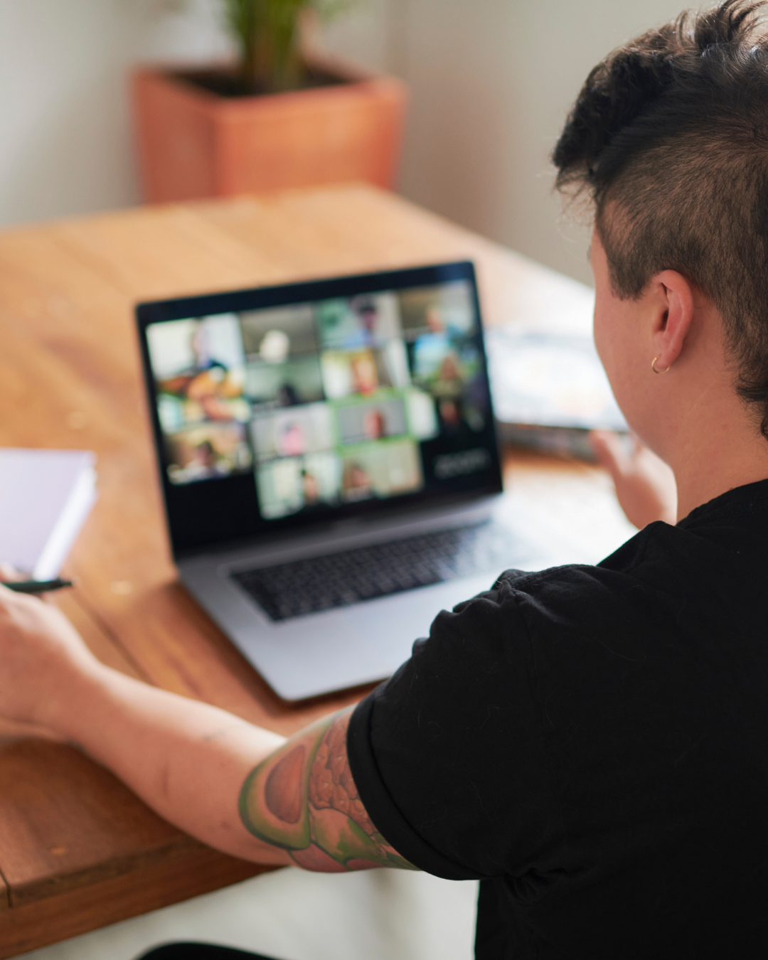 A person with tattoos on their arm participates in a virtual meeting on a laptop, with a grid of video call participants visible on the screen.