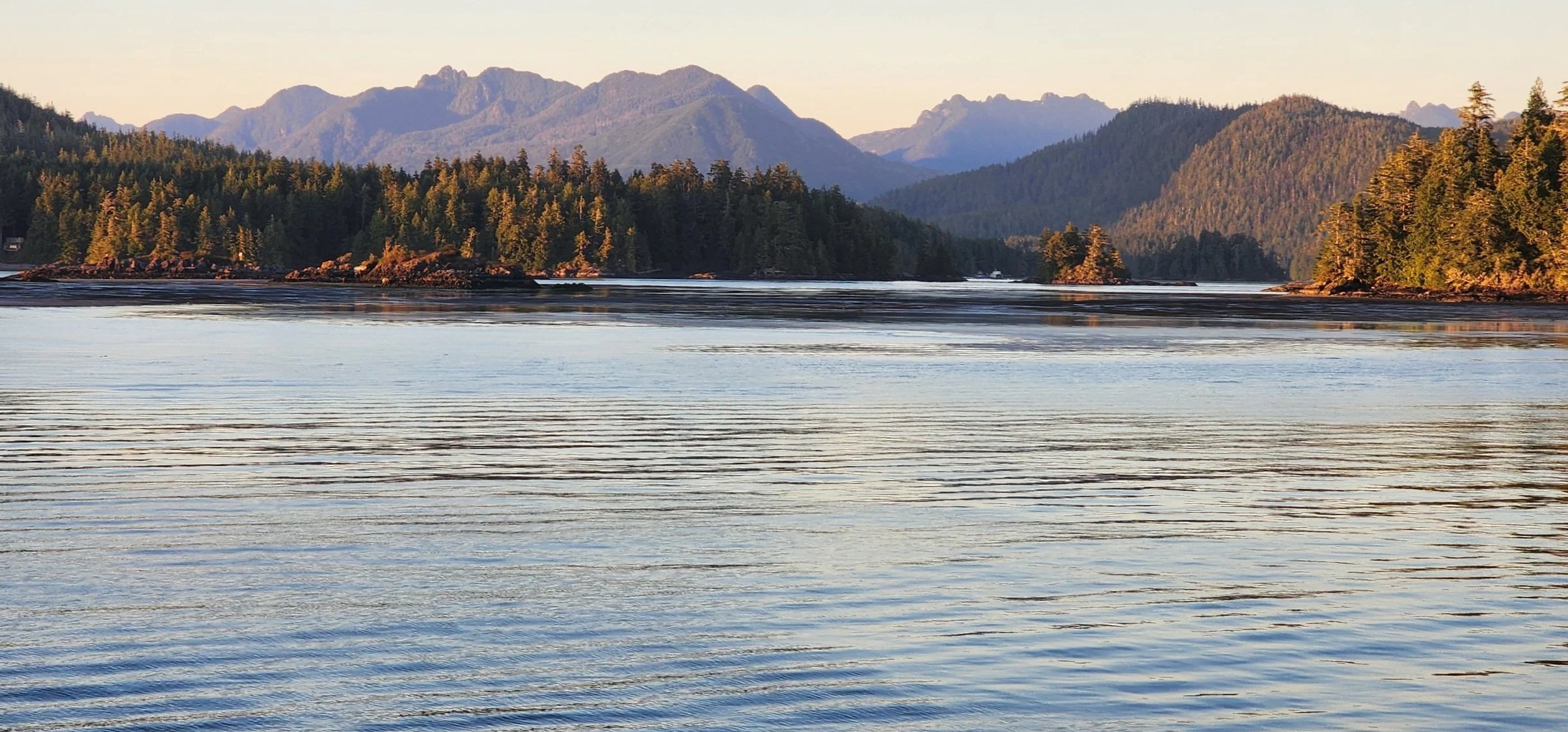 Calm river with reflections, dense evergreen forests on shorelines, and mountainous landscape in the background during sunset.