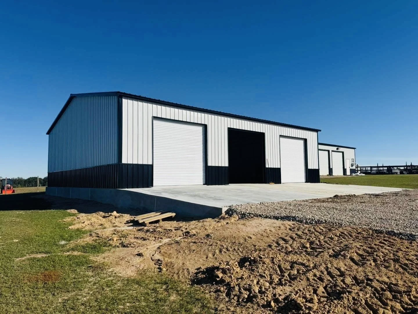 Large metal building with multiple garage doors on a clear day, surrounded by grass and dirt, with a concrete slab in front.