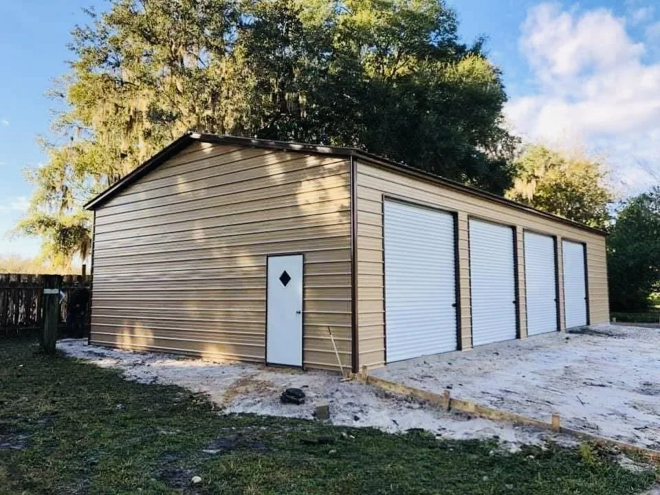 A beige metal garage with five roll-up doors and a small side door, situated on a lot with dirt and grass, surrounded by trees with a bright sky overhead.
