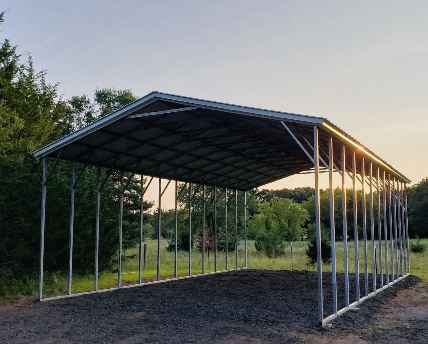 A metal building frame with a sloped roof, set outdoors during sunset, with trees and open field in the background.
