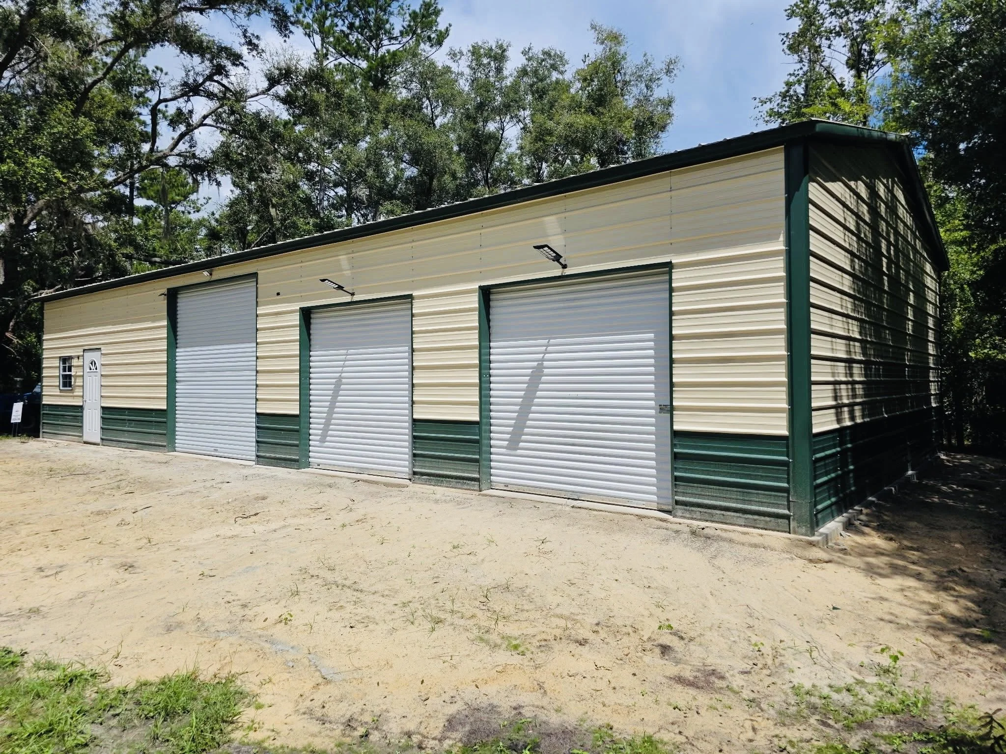 A large metal storage building with multiple roll-up doors and a white side door, surrounded by trees and on a dirt ground.