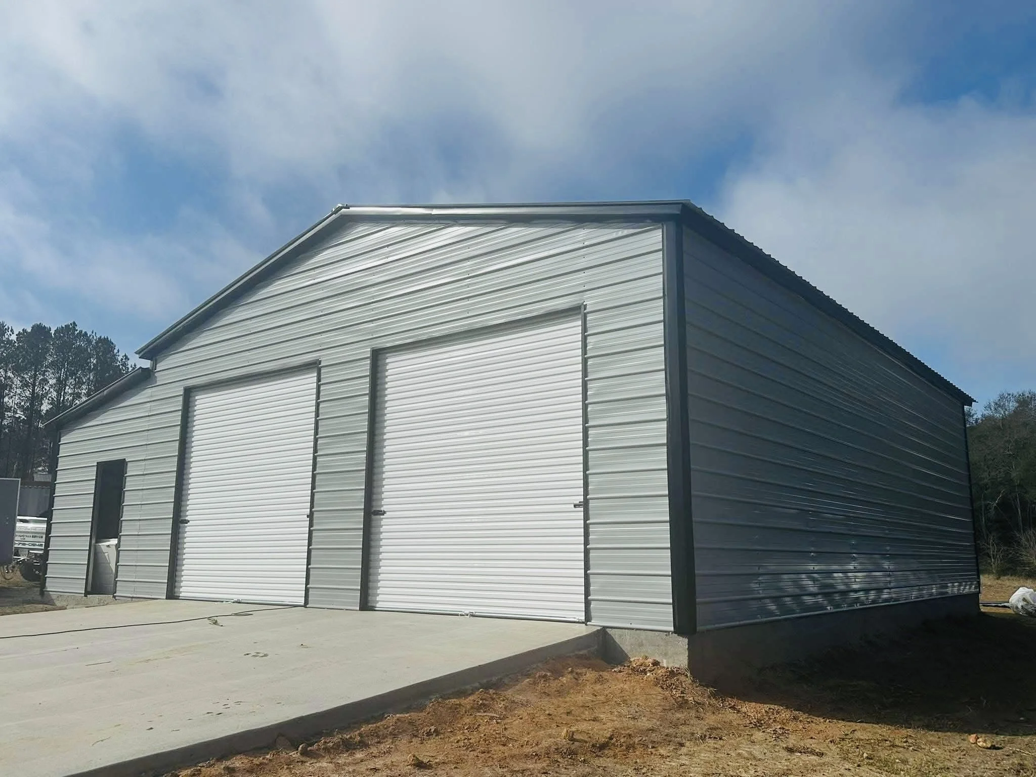The image shows the exterior of a metal building with a gable roof. The building is light-colored with closed garage doors, set against a partly cloudy sky and some trees in the background. There is a concrete driveway and a small patch of dirt in the foreground.