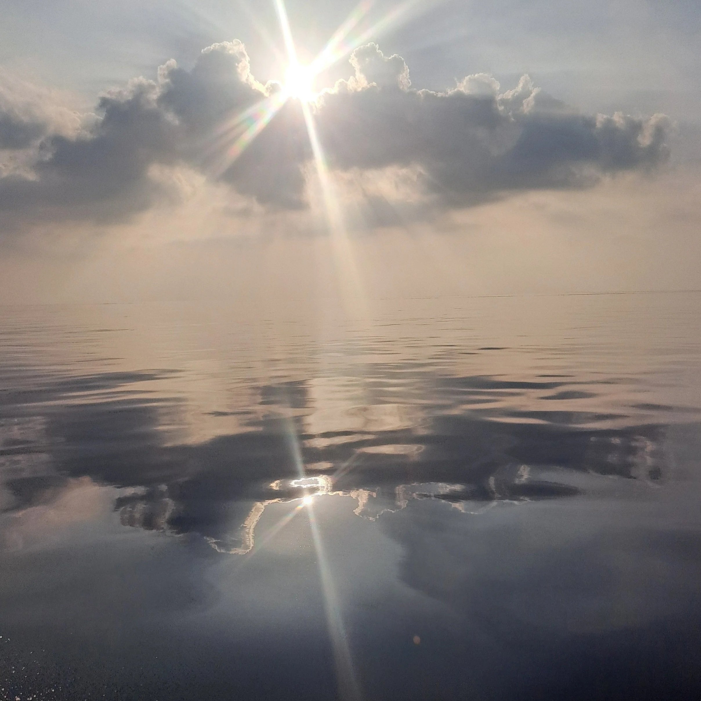 The image captures the sun shining through clouds over a large body of water, with a reflection creating a shimmering effect on the water's surface.
