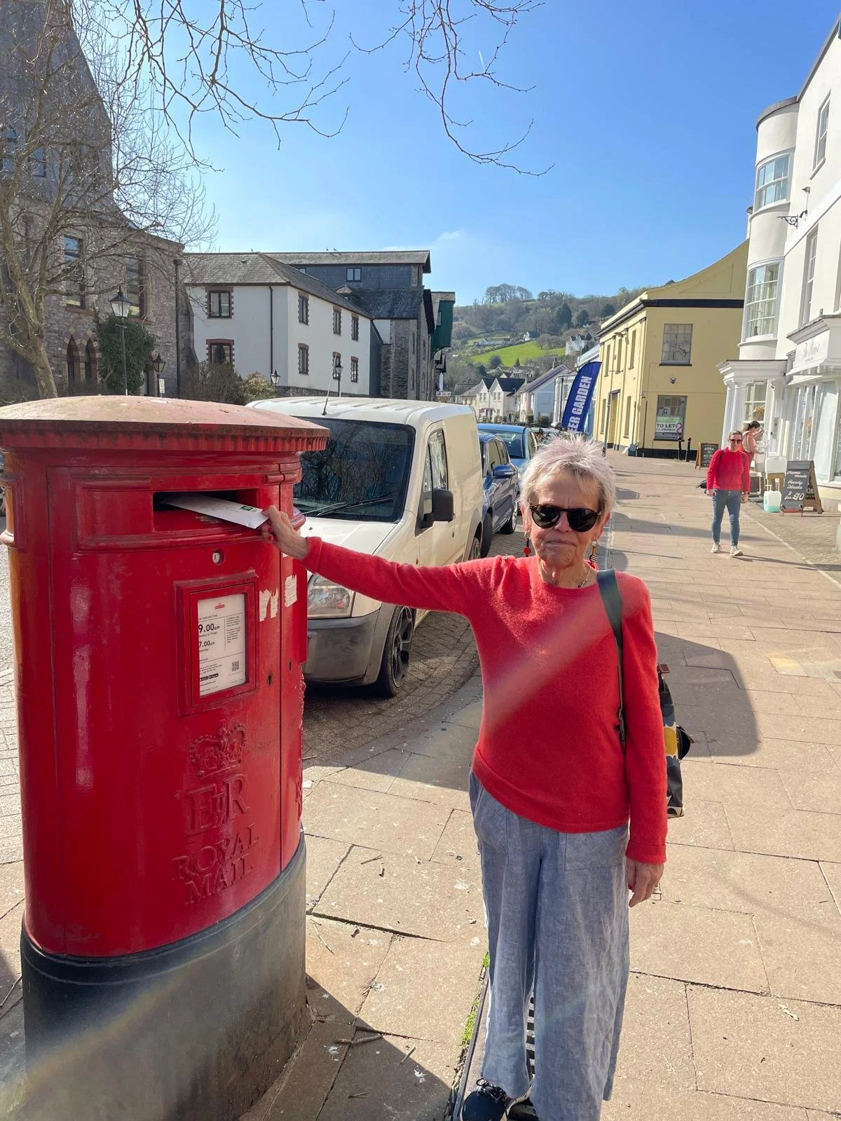 An older woman wearing sunglasses and a red sweater is mailing a letter in a red Royal Mail mailbox on a sunny street. She has short gray hair and carries a black bag over her shoulder. In the background, there are parked cars, a woman in a red jacke