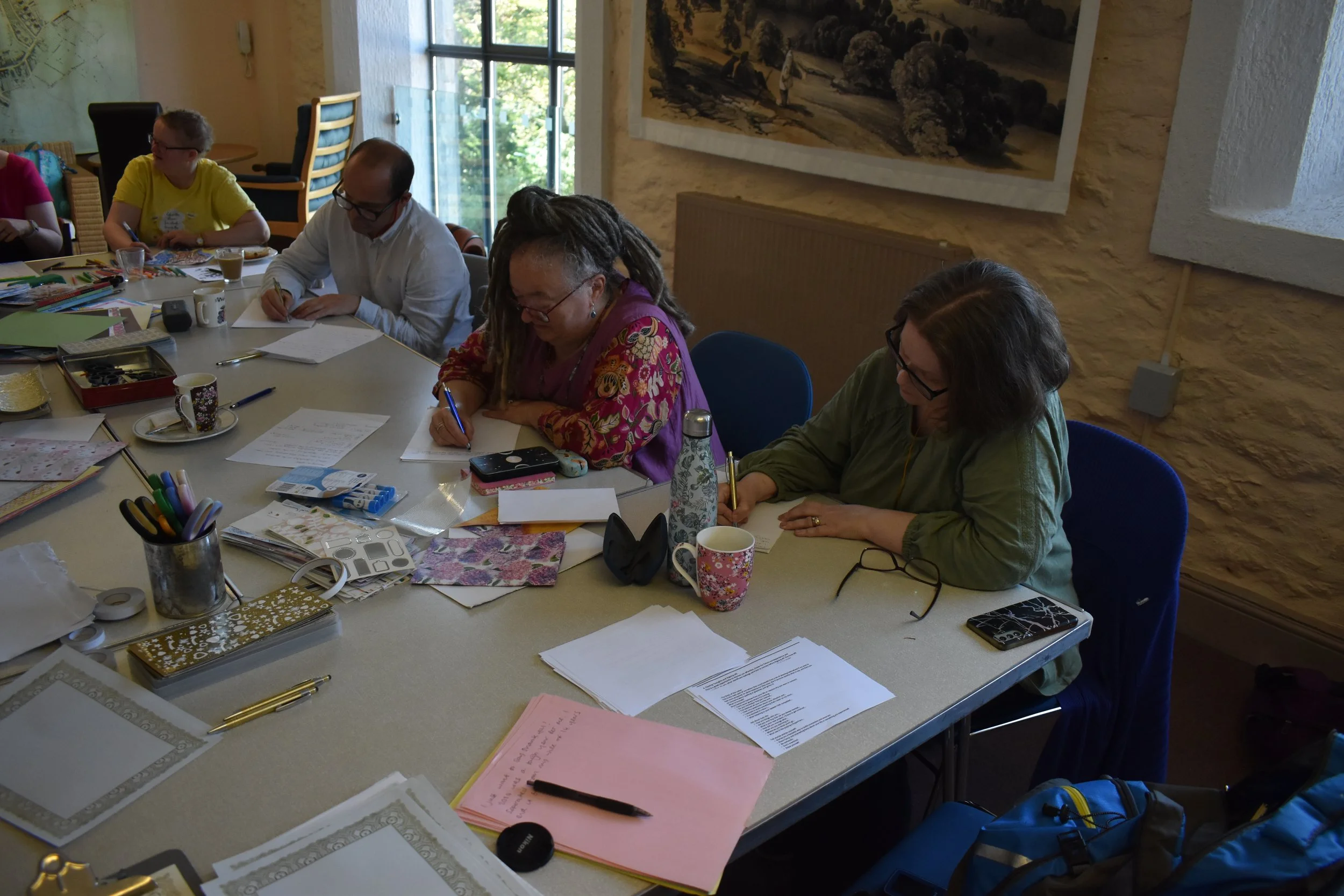 A group of five people sitting at a large table, engaged in writing or reading papers, in a room with sunlight coming through large windows and a textured stone wall decorated with artwork.