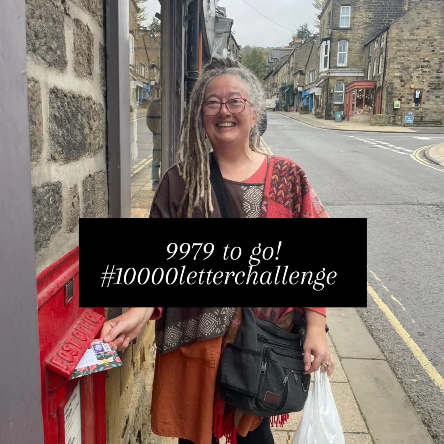A smiling woman with dreadlocks and glasses is mailing an envelope into a red postbox on a city street during daytime. The street has stone buildings and a gentle slope, with a cloudy sky overhead.