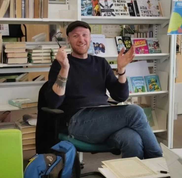 A man with a beard and tattoos sitting on a green office chair in a bookstore, smiling and holding a pen in one hand while the other hand is raised. Books and magazines are on shelves behind him, and a backpack, papers, and a laptop are on the table 