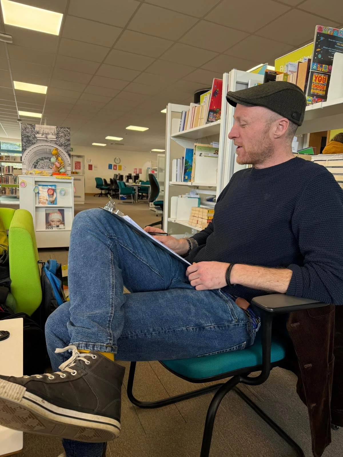 A man sitting in a library, wearing a dark sweater, blue jeans, black shoes, and a flat cap. He is reading or writing on a clipboard, with a background of bookshelves and study areas.
