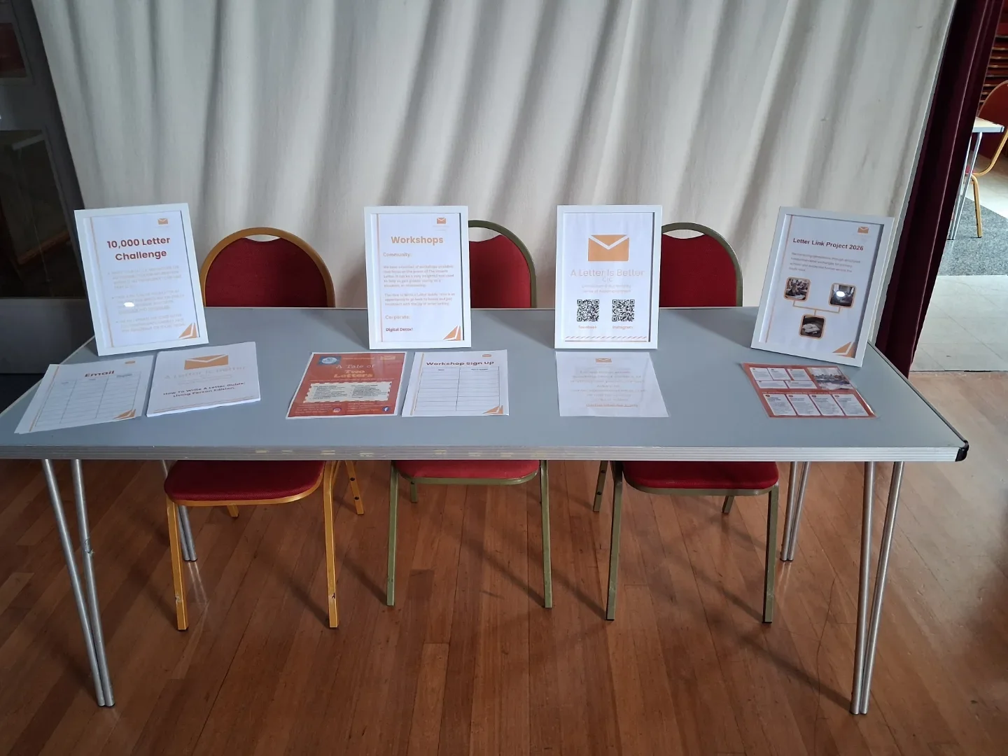Information table with posters and flyers about a letter crafting workshop, challenges, and projects, set against a white curtain background with three red chairs behind it.