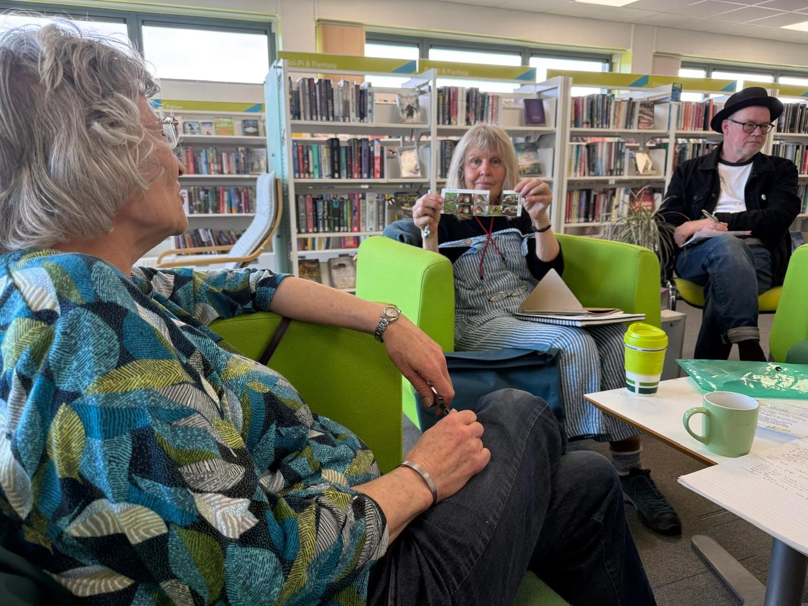 Three women and one man sitting in a library, having a conversation. One woman holds up a glass decorative item. The room has bookshelves, windows, and tables with cups and papers.