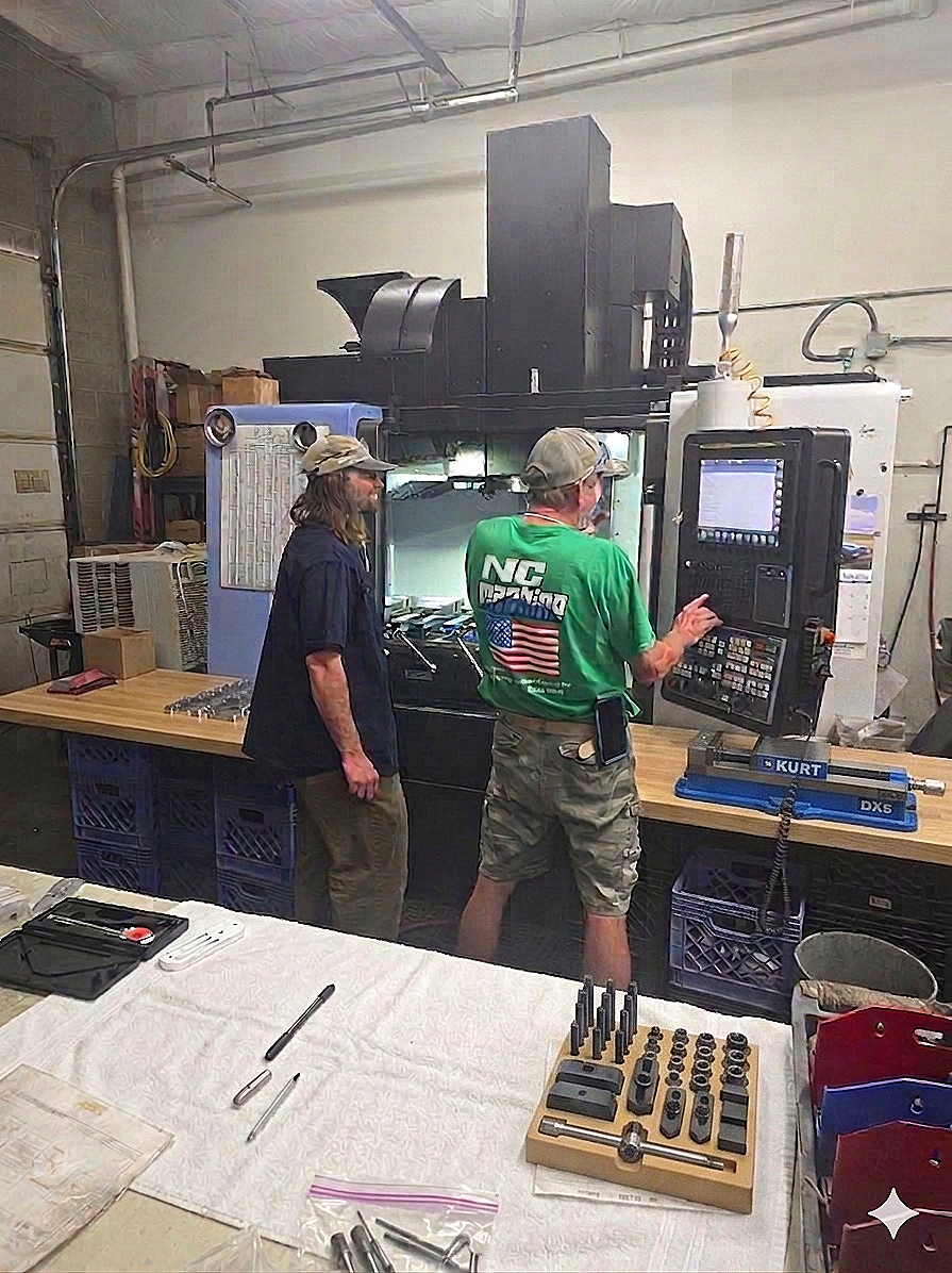 Two men working with a CNC machine in a workshop. One is operating the machine's control panel, while the other observes. The workshop is equipped with tools, parts, and safety gear.