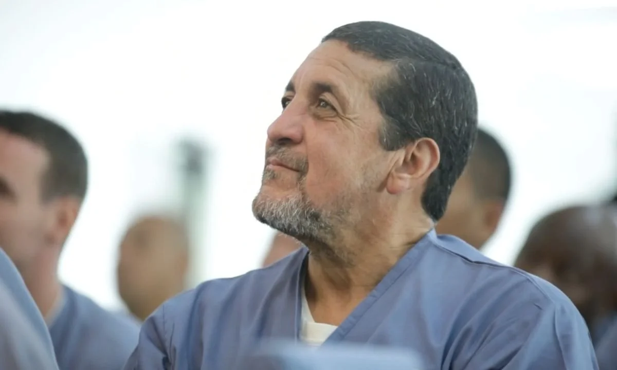 A middle-aged man with short dark hair and a beard looking up and smiling, wearing a prison uniform in a class setting with other people in similar attire in the background.