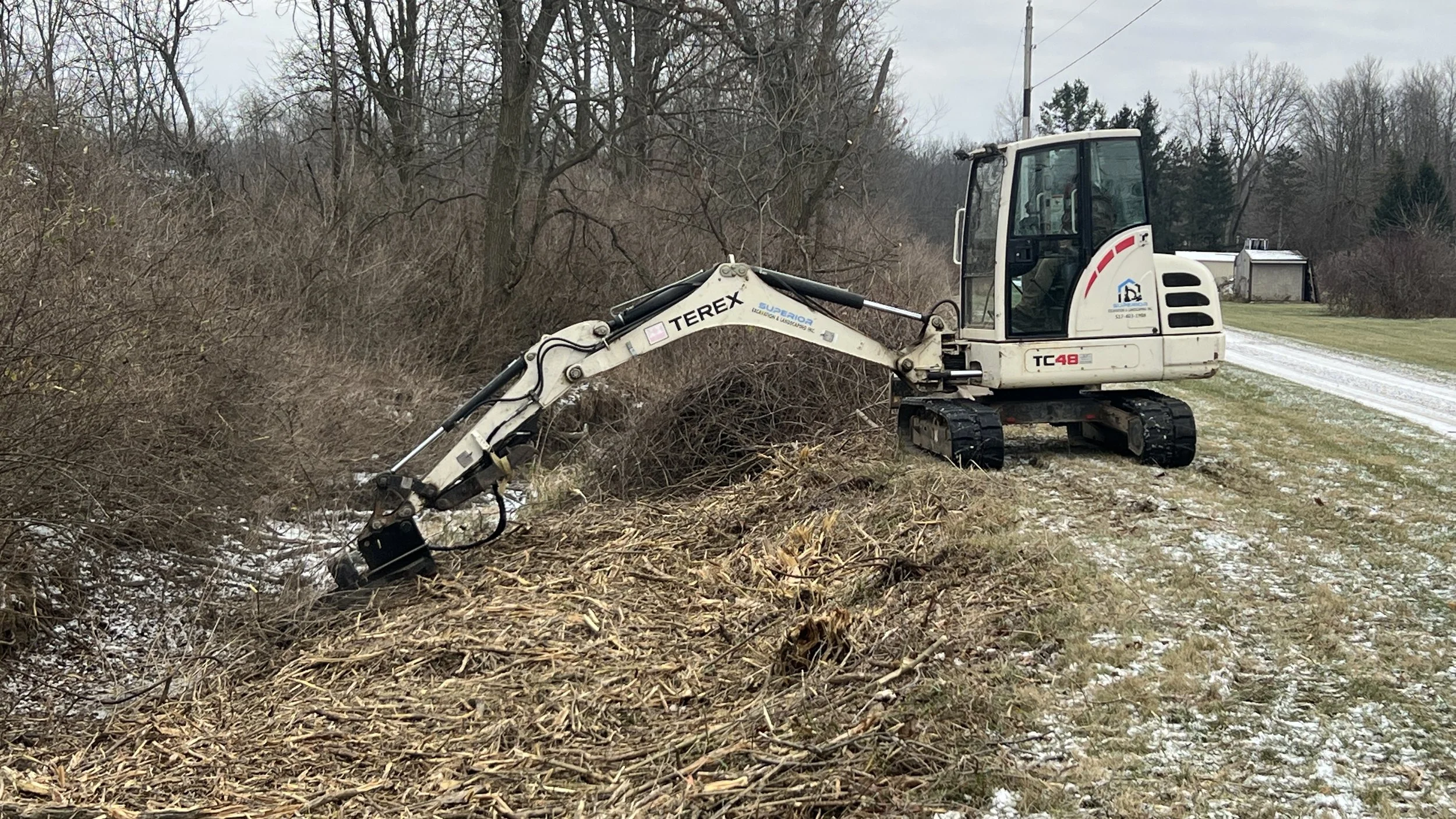 A small excavator mowing brush along the side of a rural road with some snow on the ground, trees in the background, and a small building in the distance.