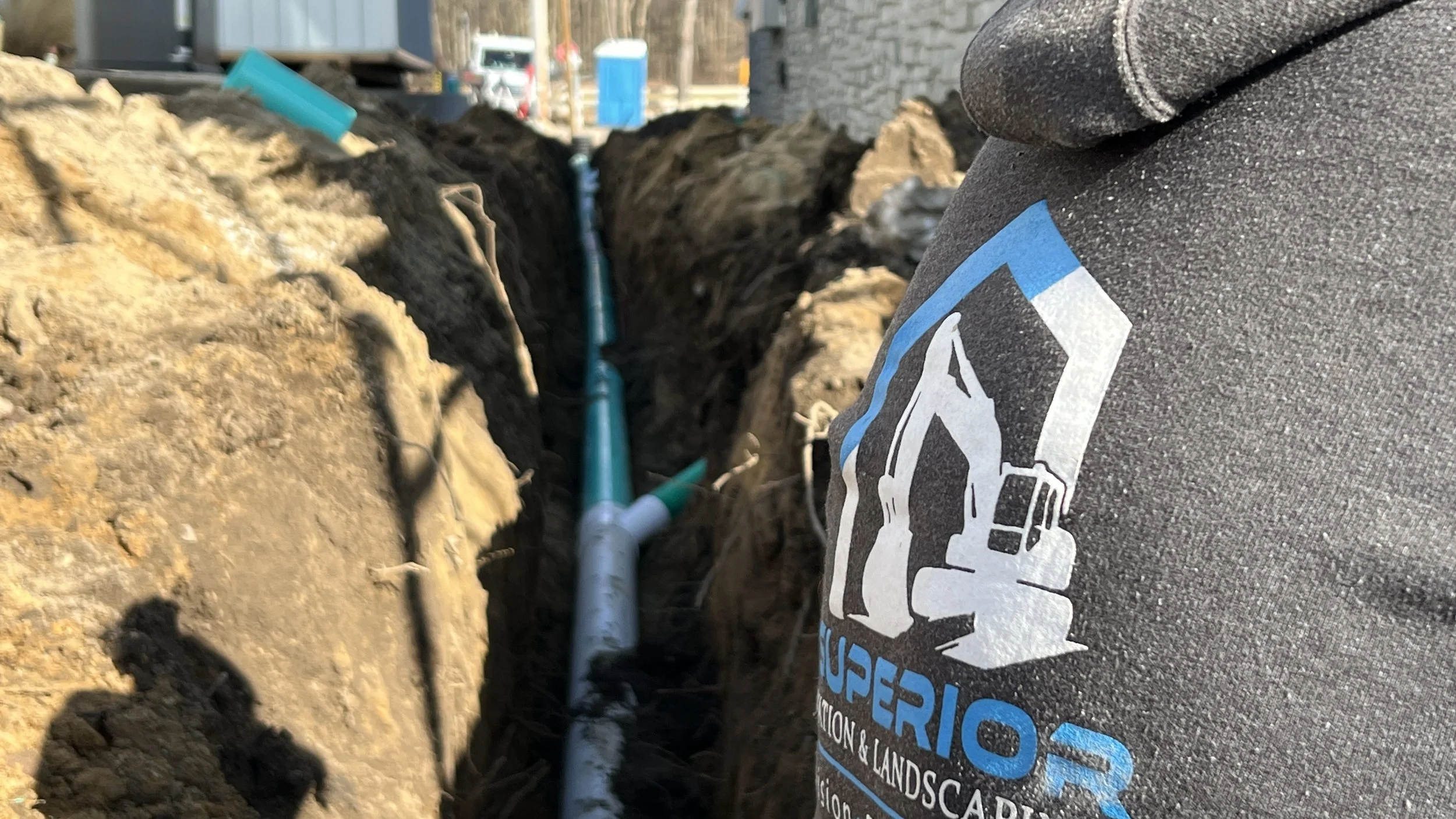 A construction worker's sleeve with a logo that says 'Superior' visible in a deep trench where blue PVC pipes are being installed. The background shows construction equipment and a blue portable toilet.