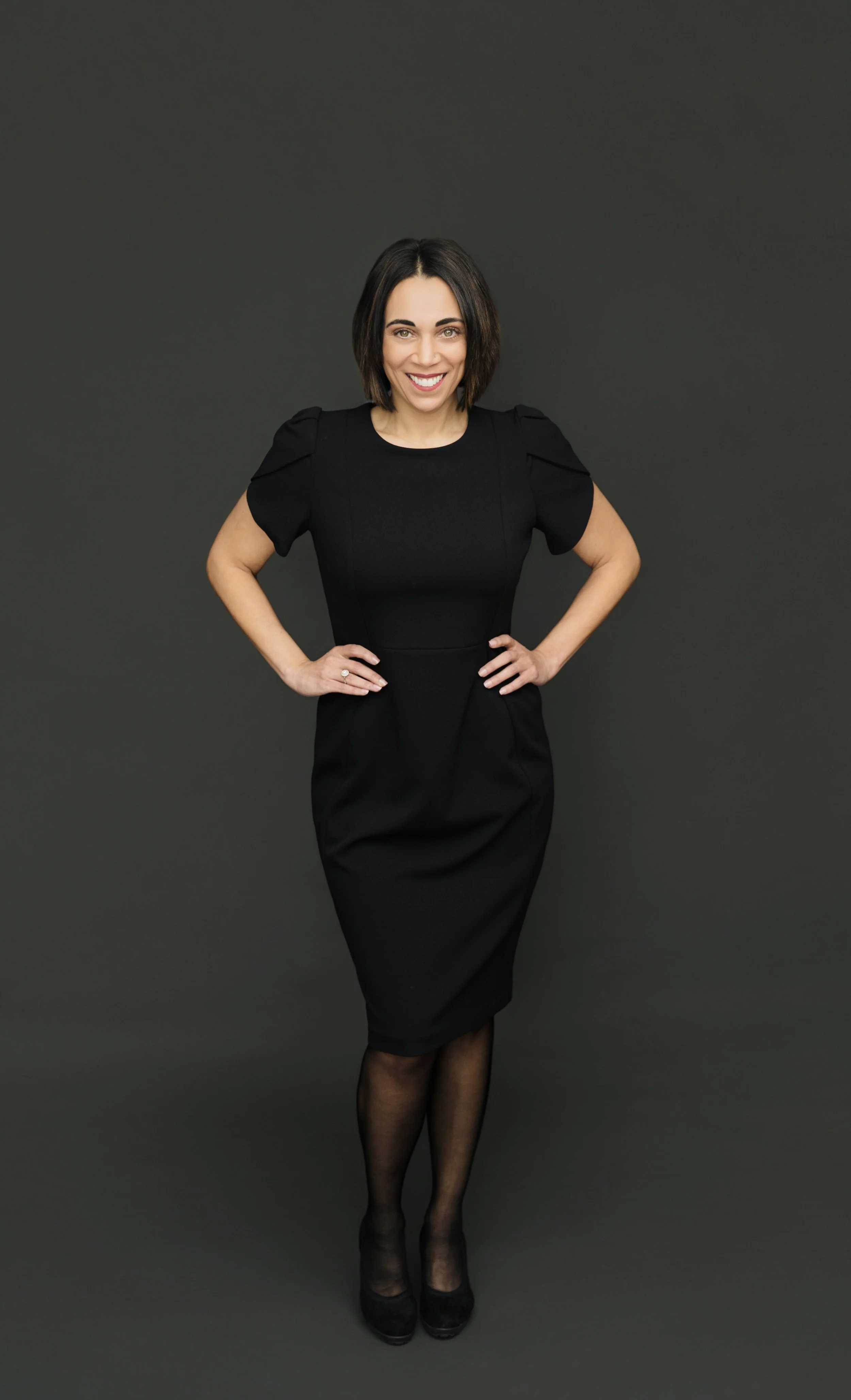 A woman smiling with hands on hips, wearing a black dress, sheer black stockings, and black heels, standing with a dark gray background.