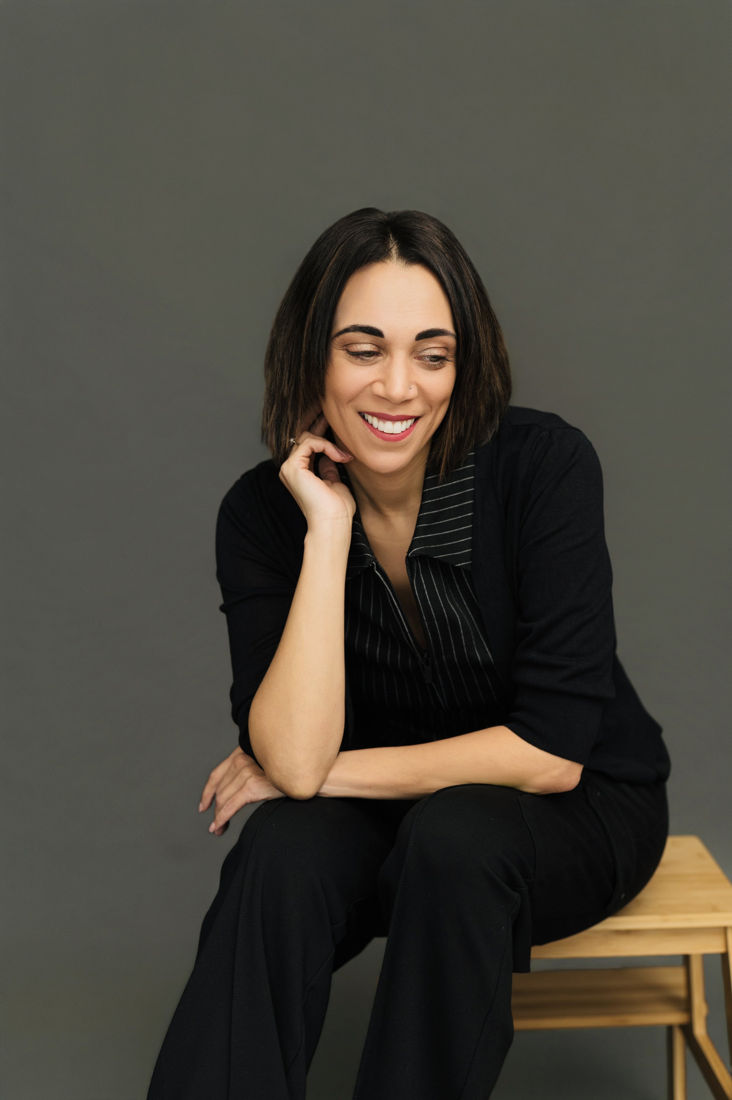 A woman with shoulder-length dark hair, smiling and looking down, sitting on a wooden bench against a dark gray background, wearing a black top and black pants.