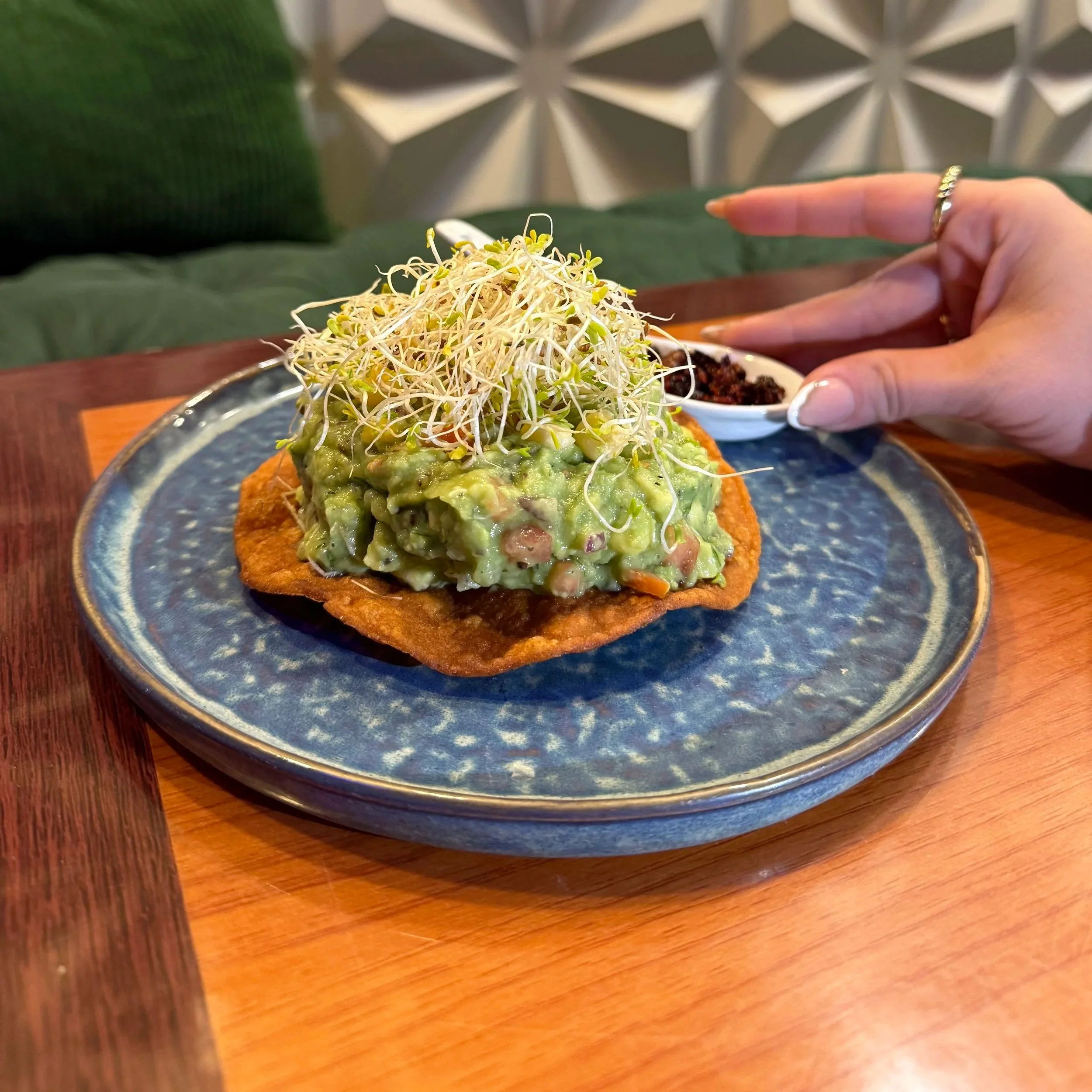 A plate of avocado toast topped with bean sprouts, served on a dark ceramic dish on a wooden table.