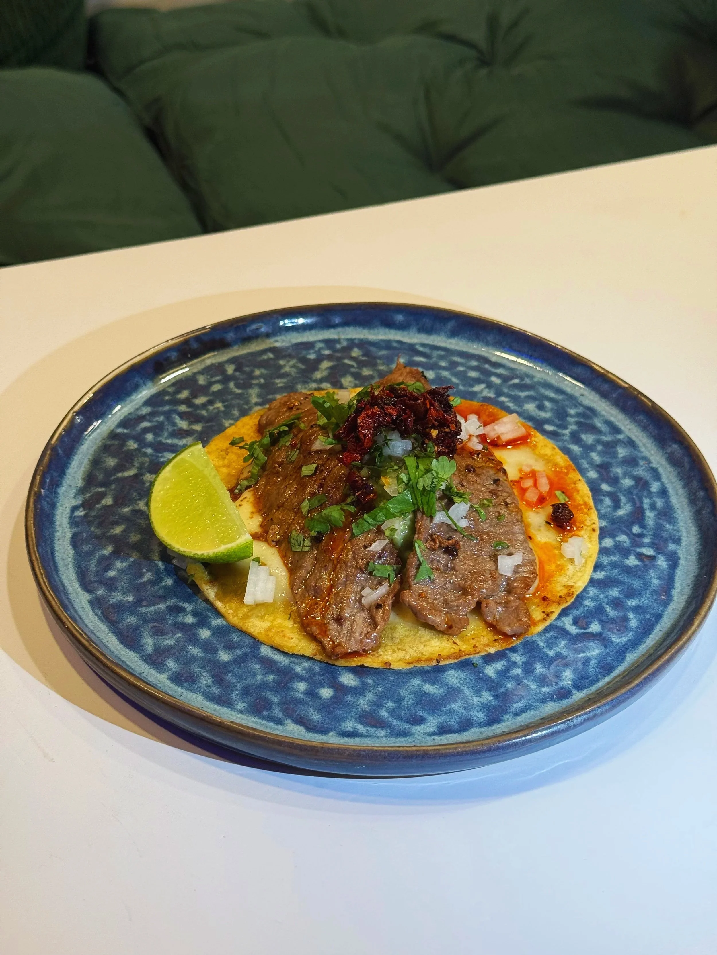 Close-up of a taco topped with cooked meat, chopped onions, cilantro, and dried red chili flakes, garnished with a lime wedge on a blue ceramic plate.