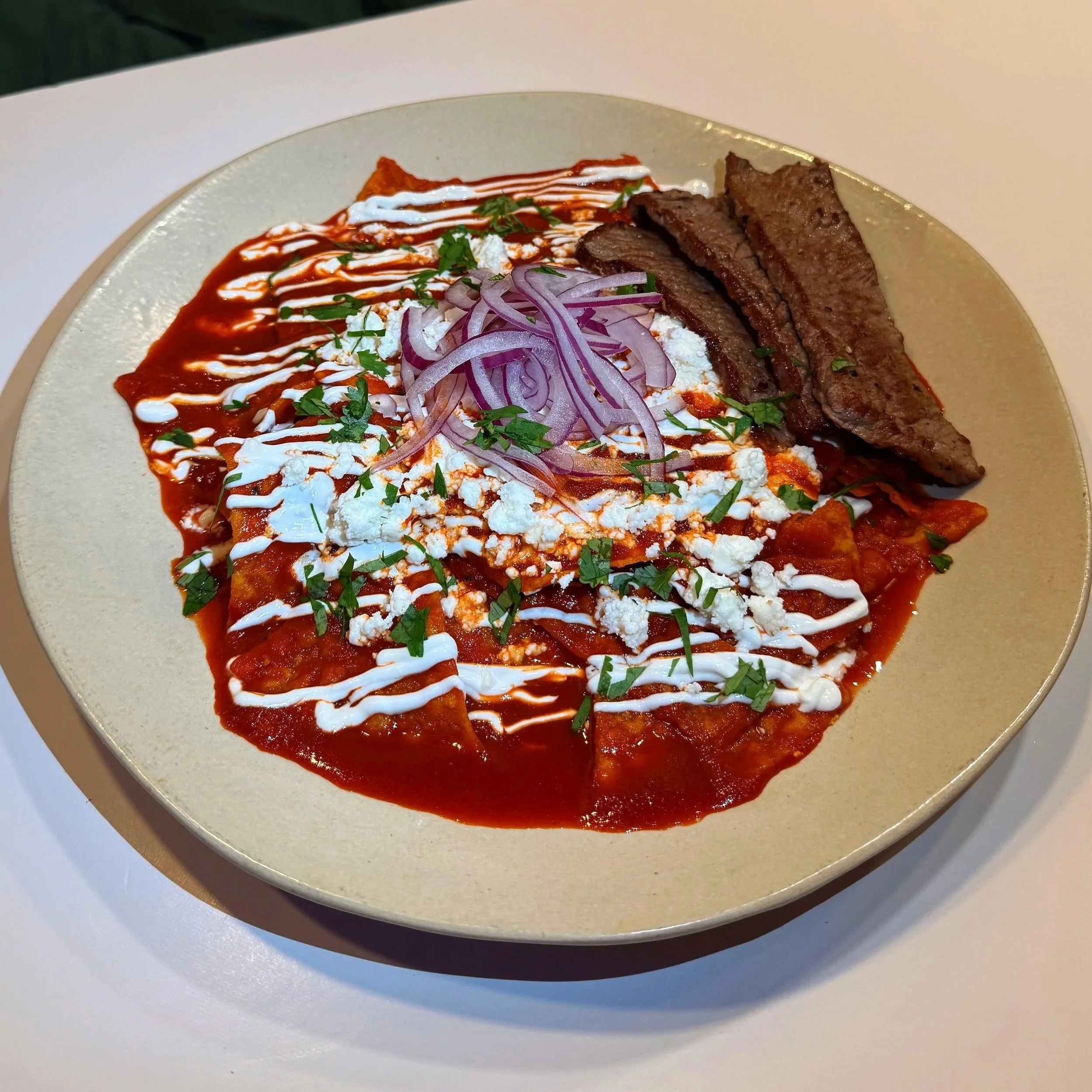 Plate of Mexican enchiladas topped with red sauce, drizzled with white crema, sprinkled with chopped cilantro, and garnished with sliced red onions.