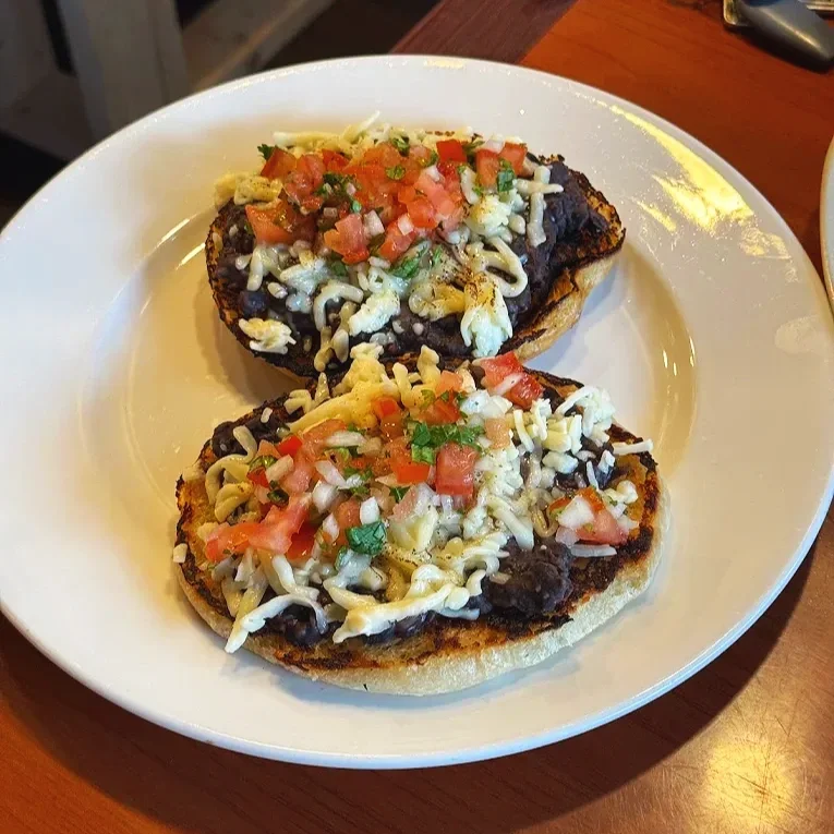 Two tostadas topped with beans, shredded cheese, diced tomatoes, and chopped cilantro on a white plate.