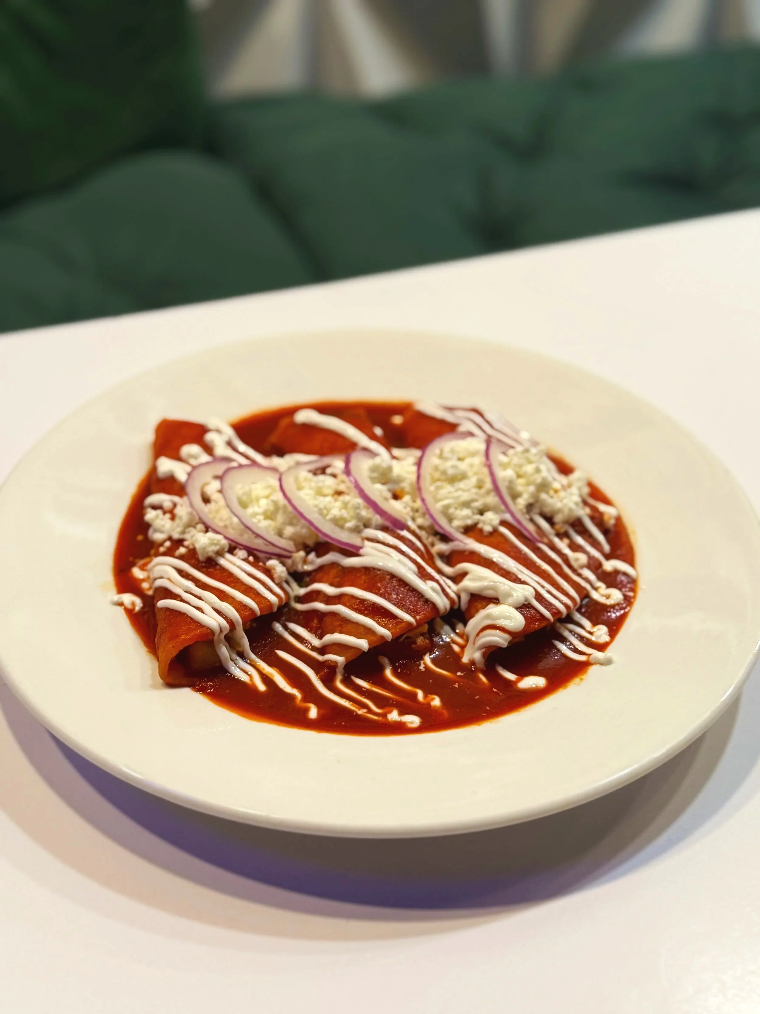 Plate of enchiladas topped with red sauce, sliced onions, crumbled cheese, and drizzled with white sauce, on a white dinner plate.