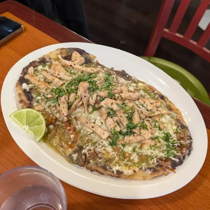 A large oval pizza topped with shredded chicken, chopped herbs, cheese, and a lime wedge on a white plate on a wooden table.